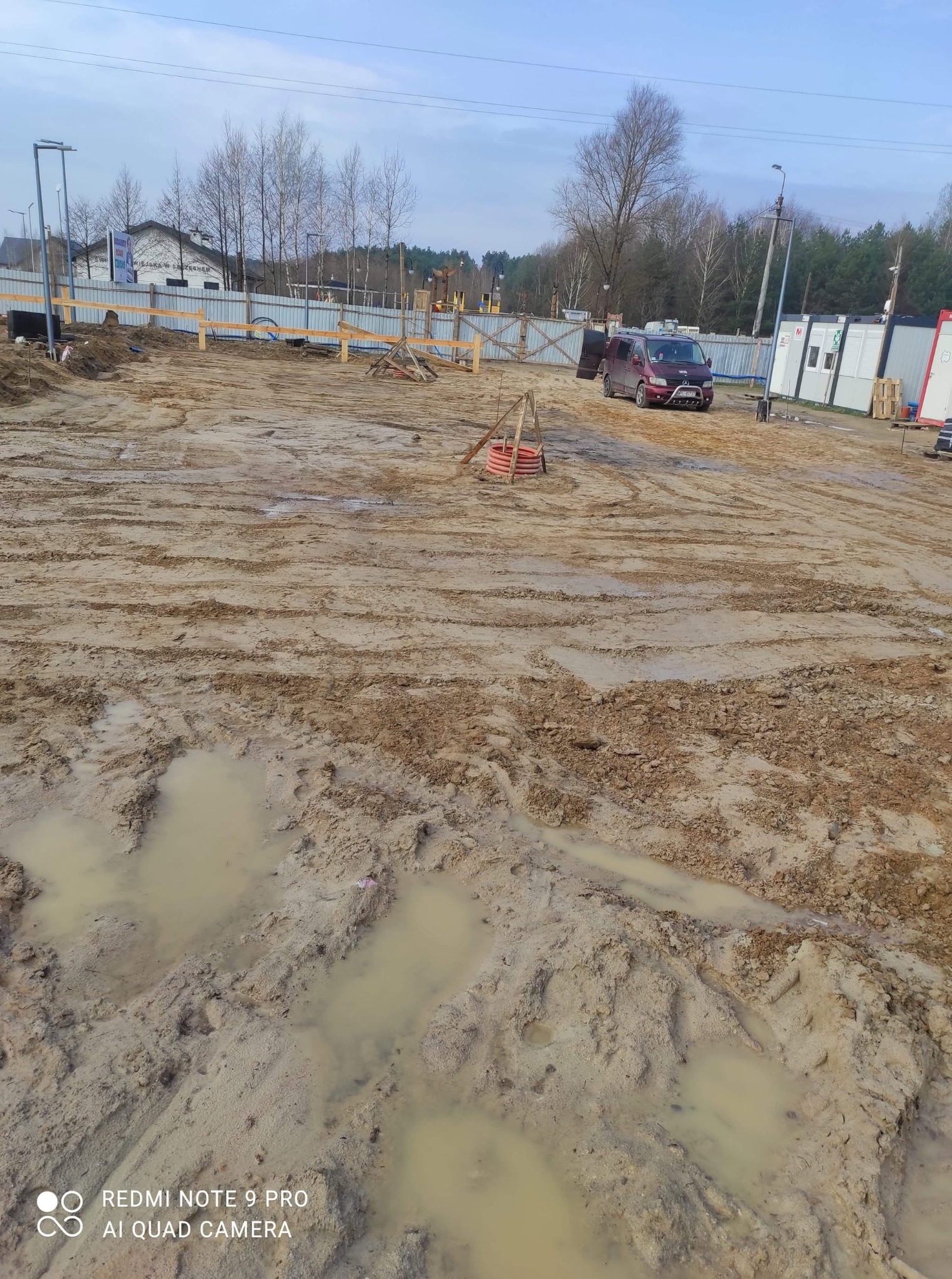 Teren budowy po deszczu: błotniste podłoże z kałużami, widoczne ślady opon, w tle ogrodzenie i budynki kontenerowe, czerwony van na uboczu.