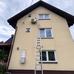 CHARUTA CHŁODNICTWO Bartosz Charuta - Żółty dom z zamontowaną klimatyzacją na ścianie. Przystawiona długa, rozłożona drabina aluminiowa. Na trawniku skrzynka z narzędziami.