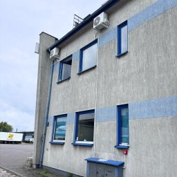 CHARUTA CHŁODNICTWO Bartosz Charuta - Szary budynek z niebieskimi akcentami i zamontowanymi klimatyzatorami na elewacji. Widoczne okna z niebieskimi ramami i skrzynka elektryczna. W tle zaparkowany samochód ciężarowy.