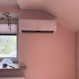 CHARUTA CHŁODNICTWO Bartosz Charuta - Biały klimatyzator Sinclair na różowej ścianie pod skosem, wyświetlający temperaturę 22 stopnie. Obok okno z roletami i zielonym bluszczem. Widoczny grzejnik.