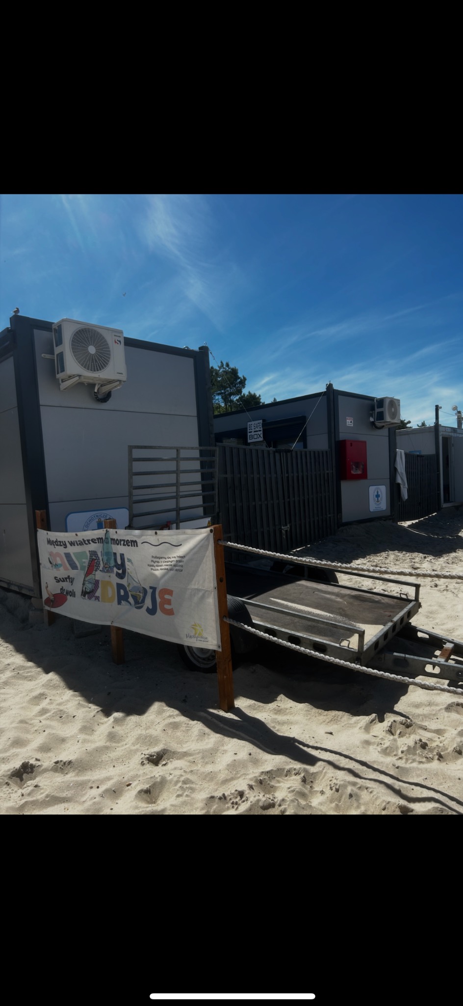 Kontenery na plaży z klimatyzacją na zewnątrz, przyczepka z banerem reklamowym. Błękitne niebo, piasek. Widok na budynki z klimatyzacją w nadmorskim otoczeniu.