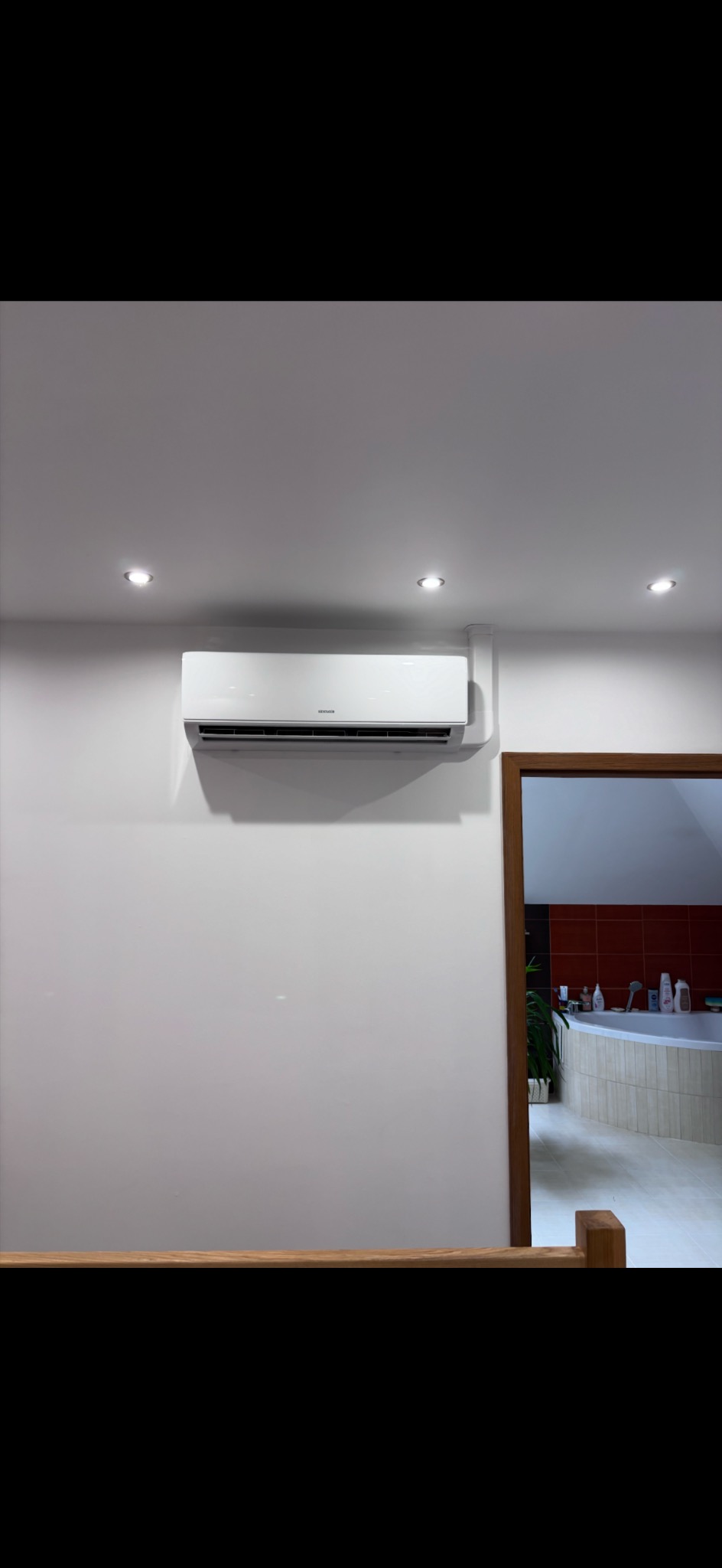 Biały klimatyzator ścienny zamontowany w jasnym pomieszczeniu z widoczną łazienką w tle. Oświetlenie punktowe sufitowe. Minimalistyczny design.