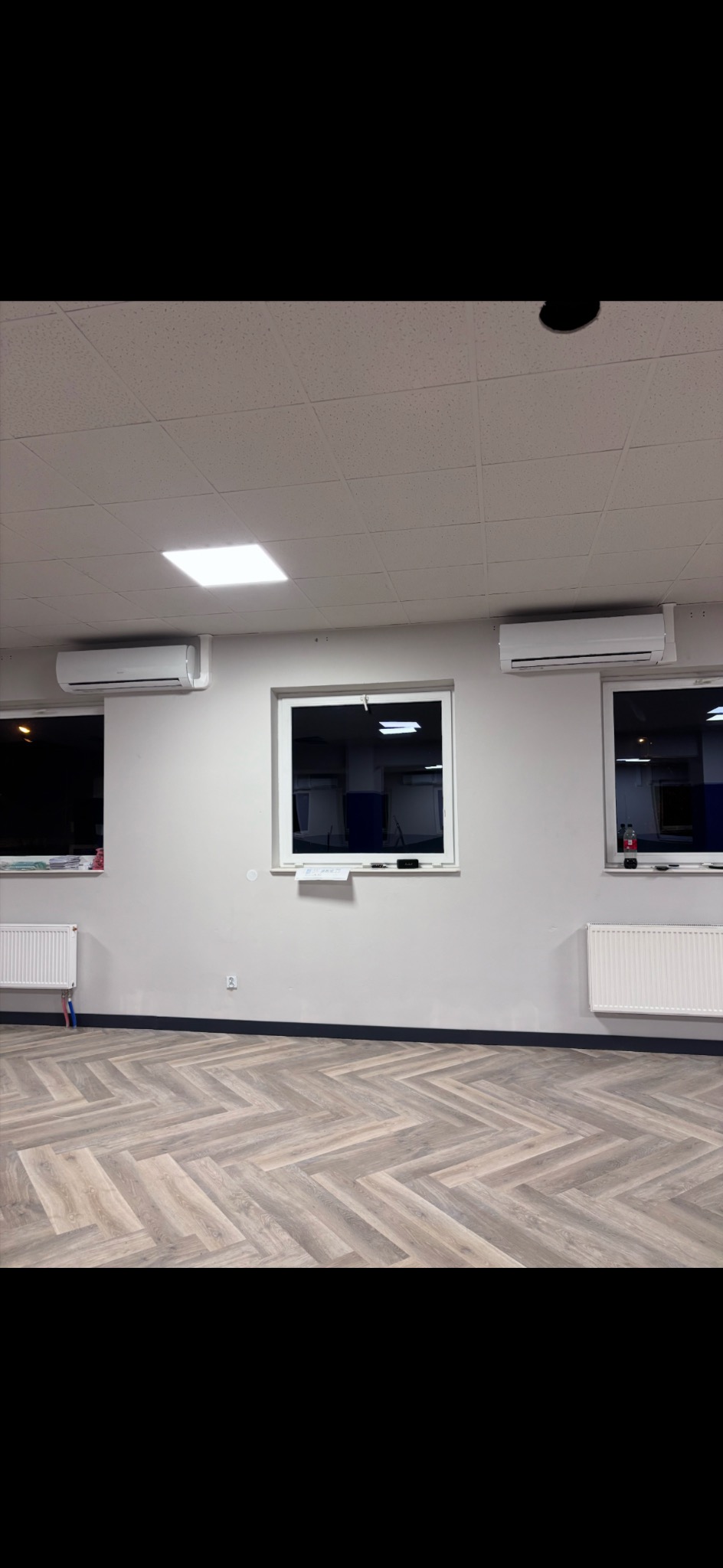 Jasne wnętrze z trzema klimatyzatorami ściennymi nad oknami i grzejnikami. Parkiet ułożony w jodełkę. Widoczne elementy instalacji elektrycznej.