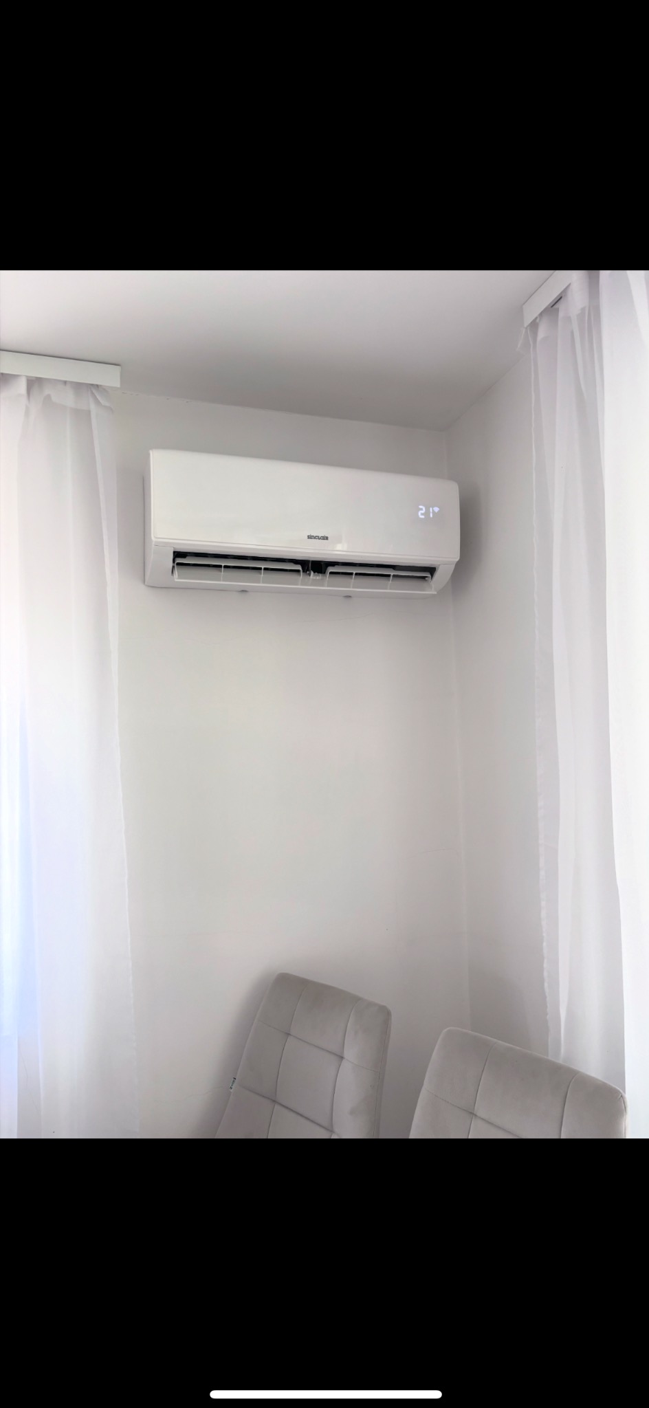 Biały klimatyzator z wyświetlaczem 21 stopni Celsjusza, zamontowany na białej ścianie w rogu pokoju, obok zwiewnych, białych zasłon i dwóch szarych krzeseł.
