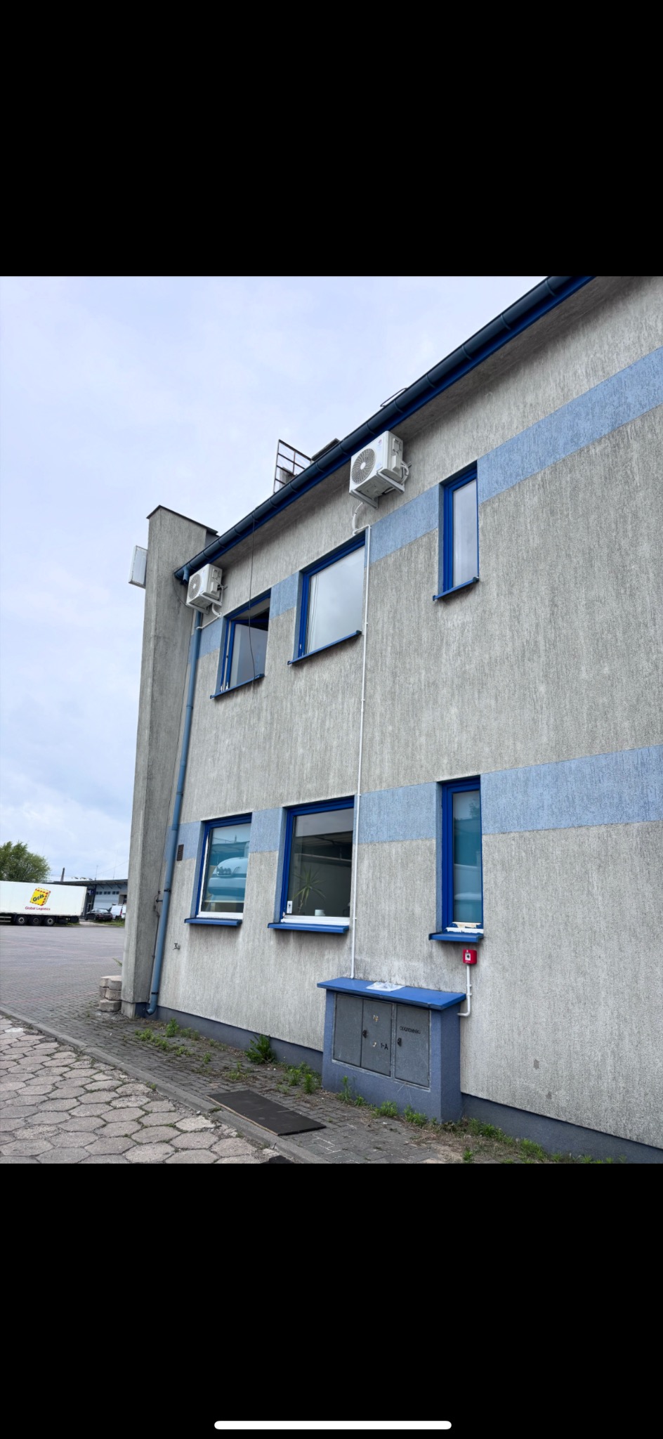 Szary budynek z niebieskimi akcentami i zamontowanymi klimatyzatorami na elewacji. Widoczne okna z niebieskimi ramami i skrzynka elektryczna. W tle zaparkowany samochód ciężarowy.