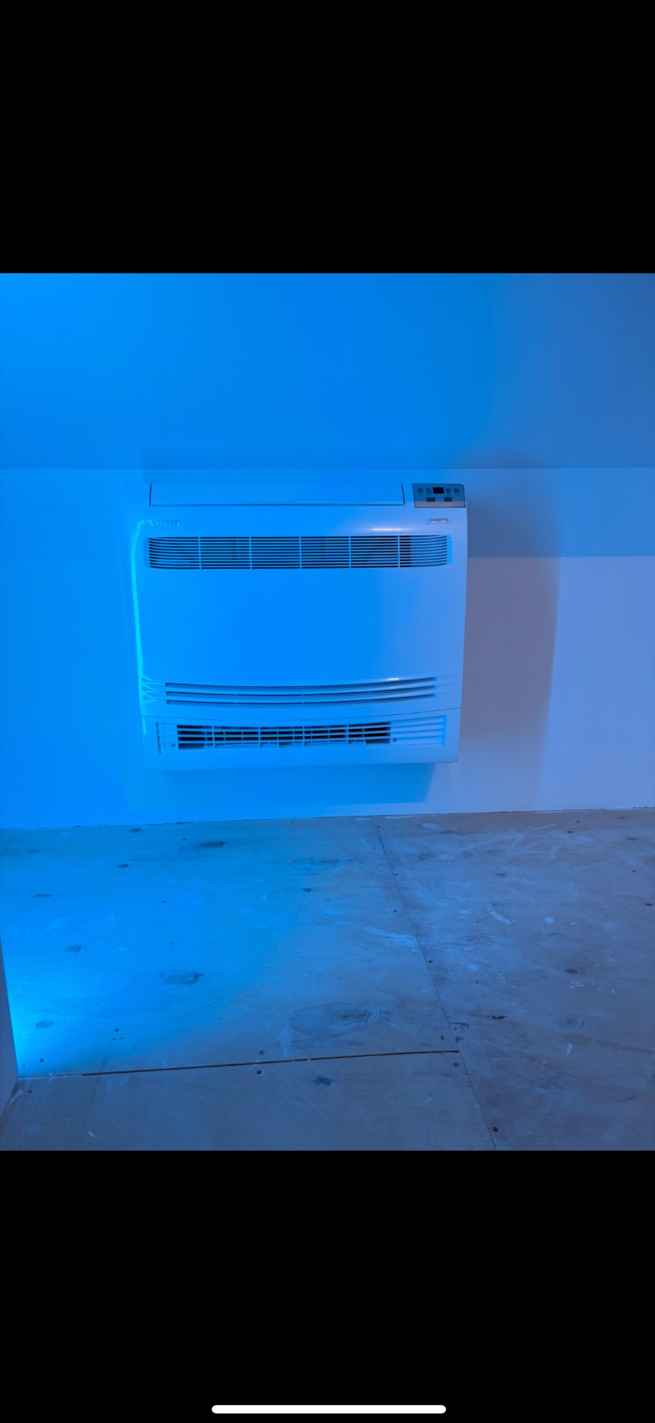 Biały klimatyzator ścienny zamontowany w pomieszczeniu z surową podłogą, oświetlony niebieskim światłem. Widoczne detale urządzenia i fragment podłogi.