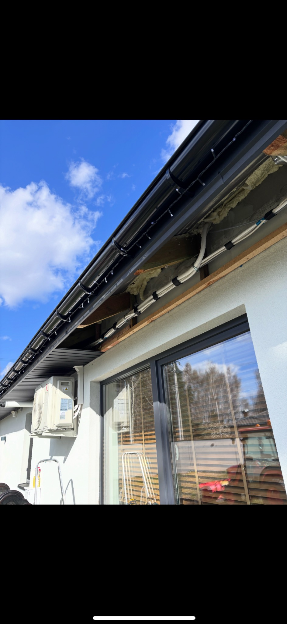 Biała jednostka klimatyzacji zewnętrznej zamontowana na ścianie domu, widoczne rury i instalacja pod dachem, błękitne niebo w tle.