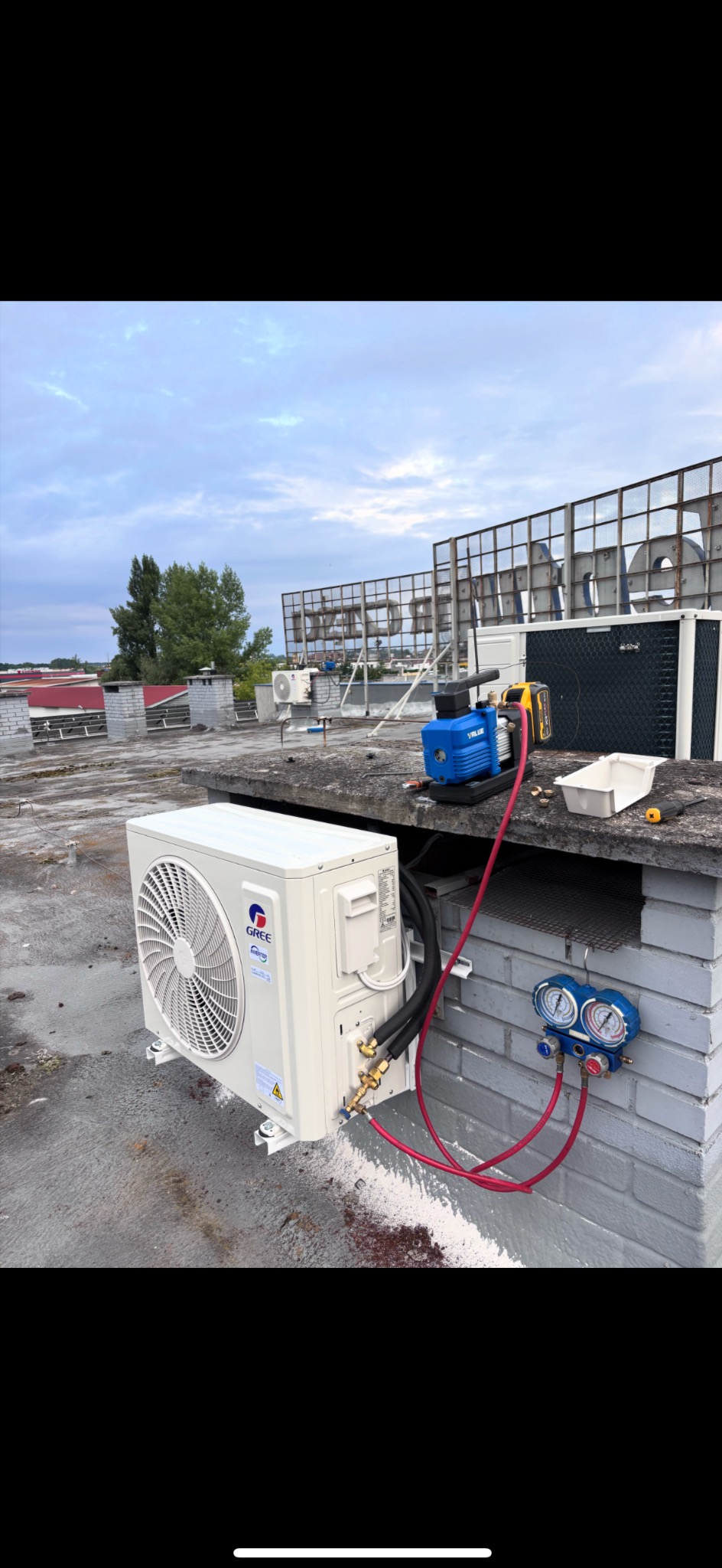 Biały klimatyzator Gree na dachu budynku, podłączony do niebieskiej pompy próżniowej i manometru. Widoczne przewody i narzędzia. W tle inne urządzenia i konstrukcja.