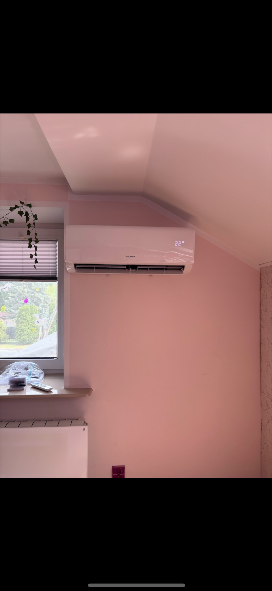 Biały klimatyzator Sinclair na różowej ścianie pod skosem, wyświetlający temperaturę 22 stopnie. Obok okno z roletami i zielonym bluszczem. Widoczny grzejnik.