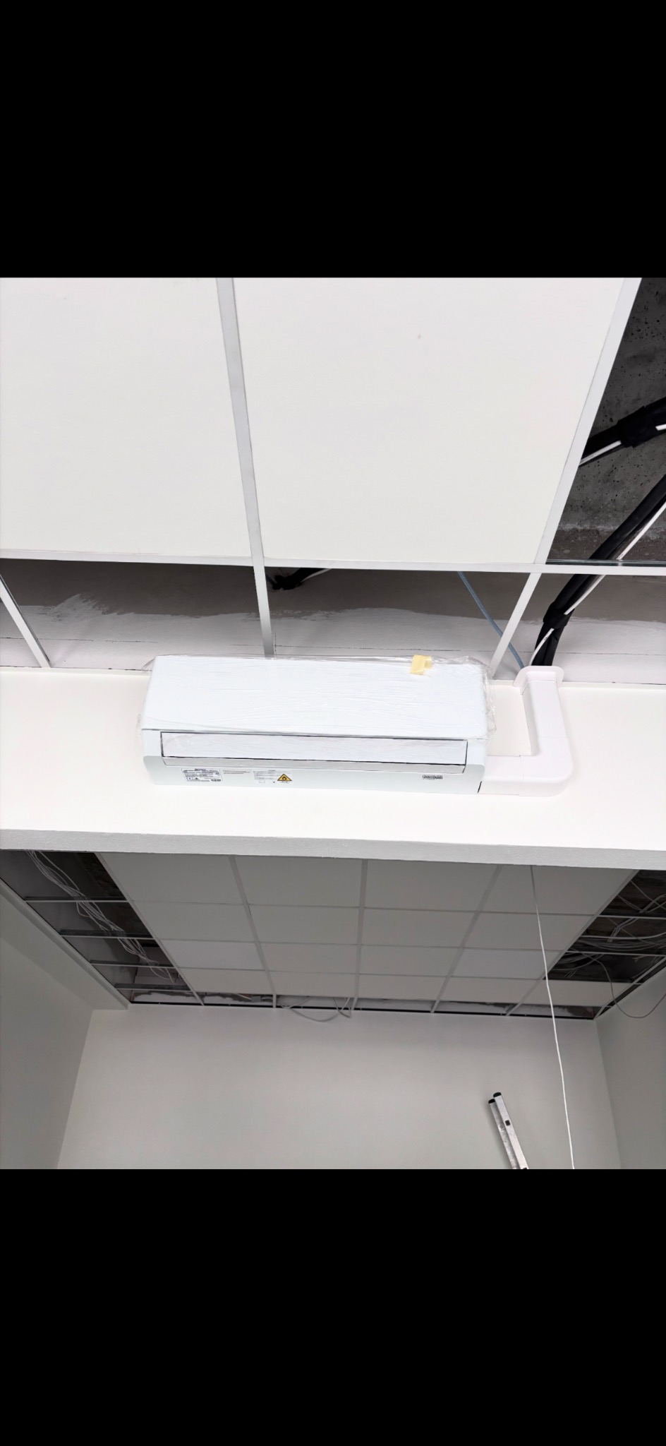 Biały klimatyzator ścienny zamontowany pod sufitem kasetonowym w jasnym pomieszczeniu. Widoczne elementy instalacji i drabina. Częściowo folia ochronna na urządzeniu.