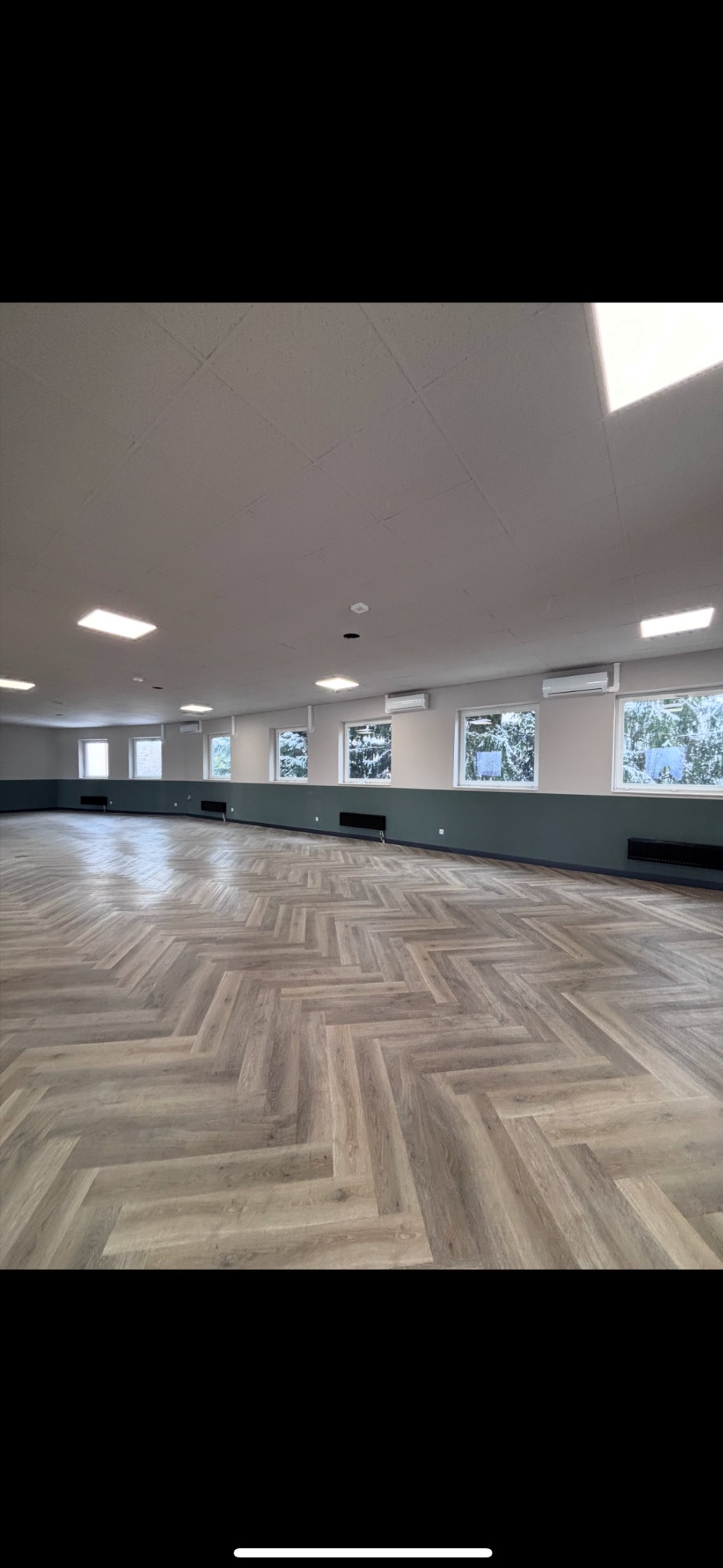 Jasne, przestronne wnętrze z parkietem w jodełkę, klimatyzatorami pod oknami i ciemnozieloną lamperią. Widok na zasypany śniegiem krajobraz za oknami.