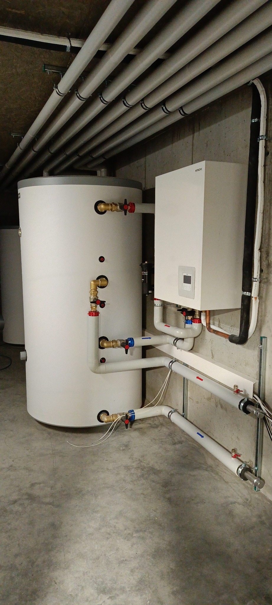 Biały zasobnik ciepłej wody i wiszący piec gazowy Hitachi z rurami w piwnicy. Instalacja z zaworami i rurami na betonowej ścianie i podłodze. Rury pod sufitem.
