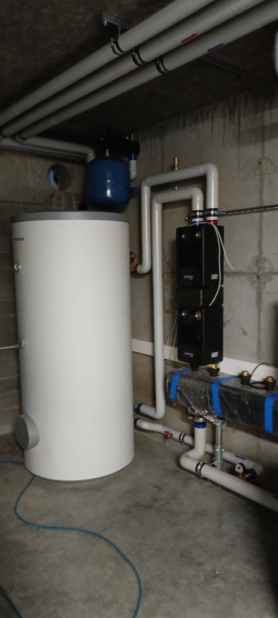 Biały zasobnik ciepłej wody z niebieskim zbiornikiem wyrównawczym, podłączony do instalacji z szarych rur w surowym pomieszczeniu. Widoczne czarne moduły sterujące.