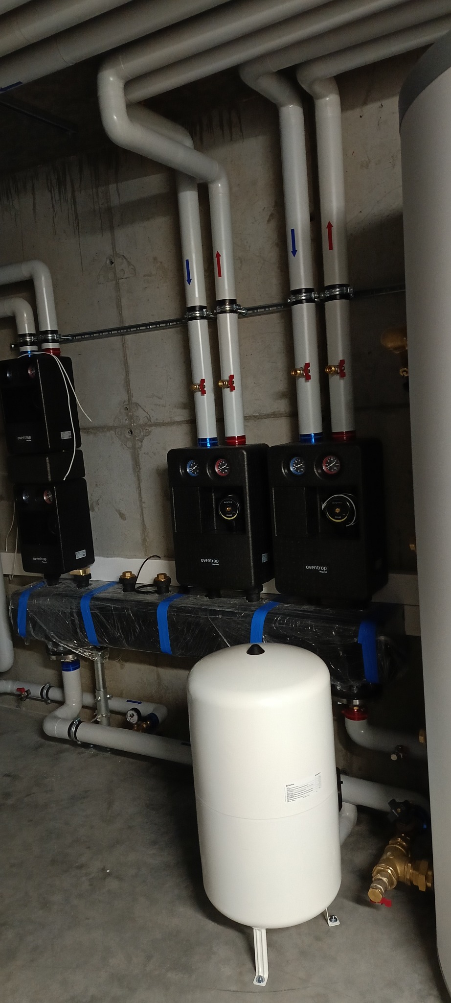 Złożona instalacja grzewcza z czarnymi modułami Eventrop, białym zbiornikiem wyrównawczym i rurami z oznaczeniami przepływu (czerwone i niebieskie strzałki).