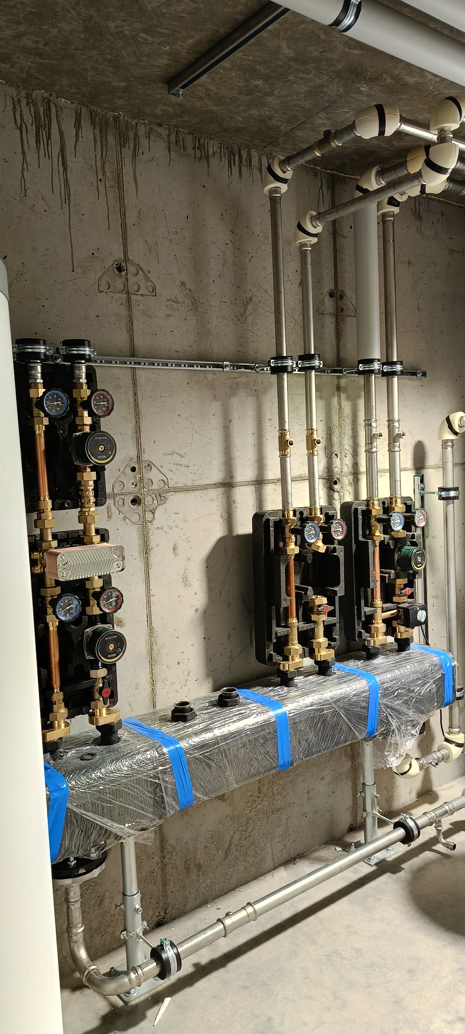Złożona instalacja grzewcza z rurami miedzianymi i stalowymi, manometrami i pompami obiegowymi na tle surowej, betonowej ściany. Elementy zabezpieczone folią i taśmą.