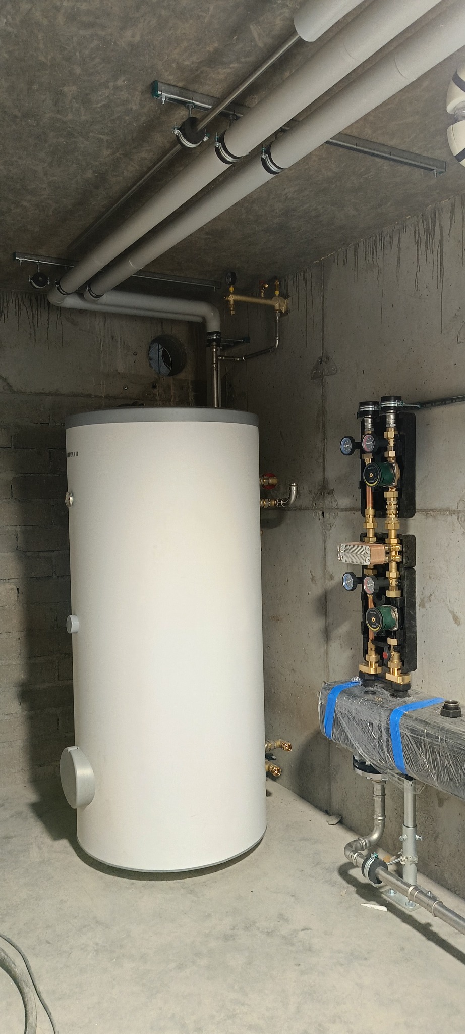 Biały bojler z podłączeniami w surowym pomieszczeniu. Widoczne rury pod sufitem i elementy instalacji na ścianie. Ujęcie z dołu, podkreślające wysokość instalacji.