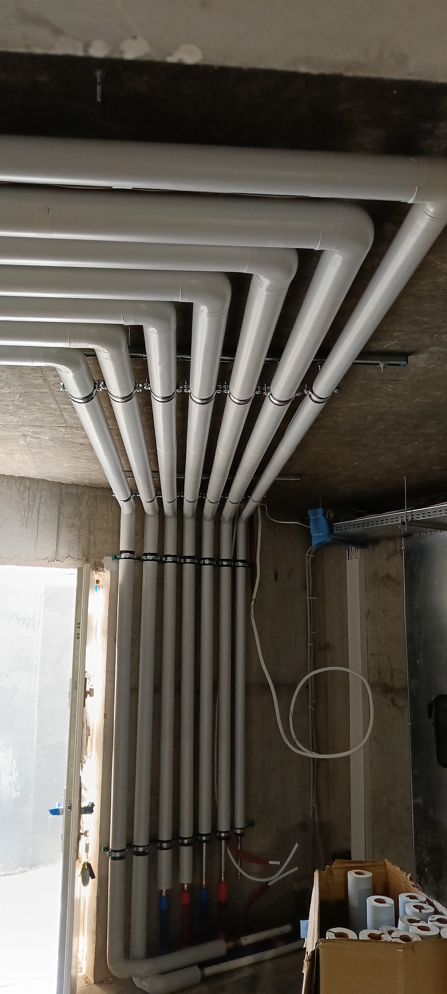 Białe rury instalacyjne równolegle ułożone pod betonowym sufitem, przymocowane metalowymi obejmami. Widoczne elementy instalacji i fragment ściany z oknem.