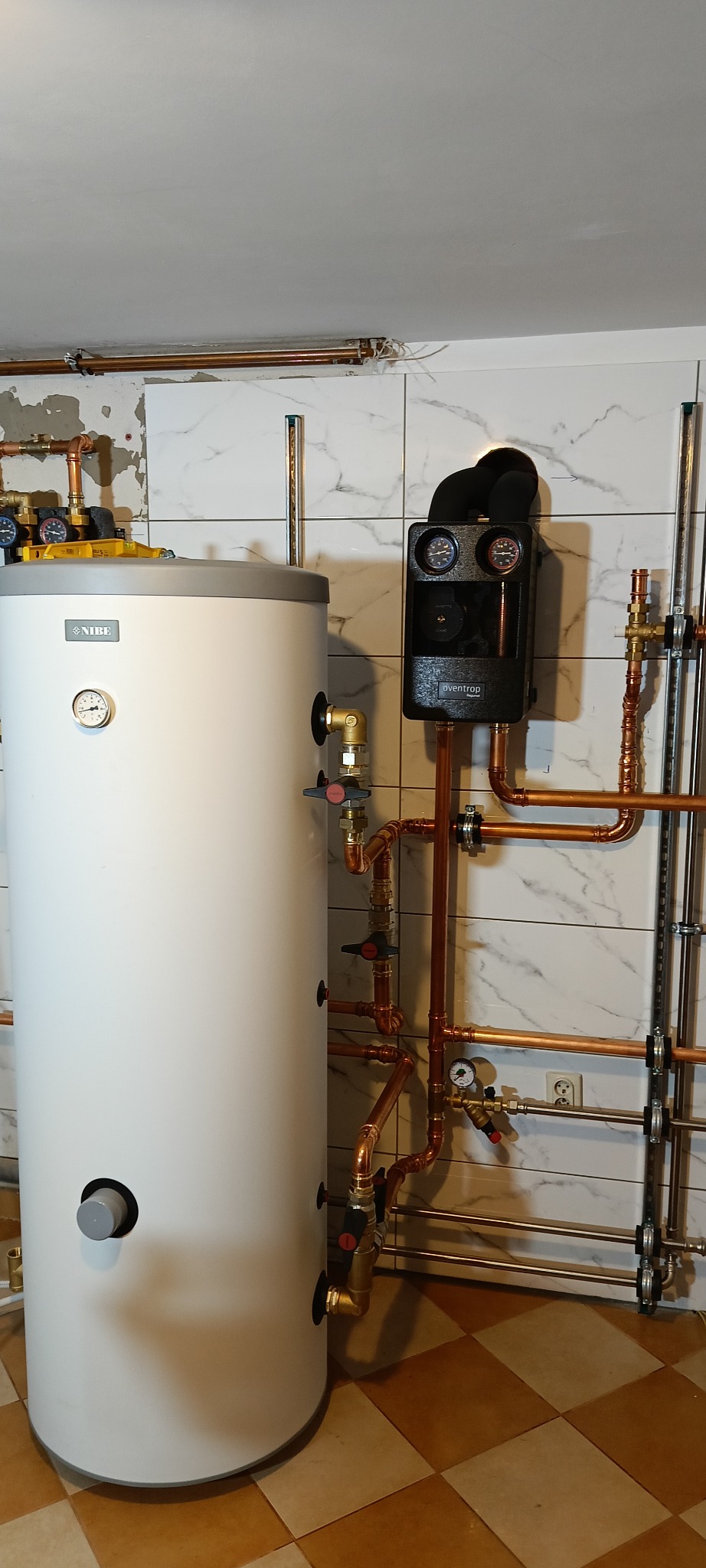 Biały bojler Nibe z czarnym regulatorem Oventrop i miedzianą instalacją. Widoczne liczne zawory i manometry. Tło stanowią białe płytki imitujące marmur.