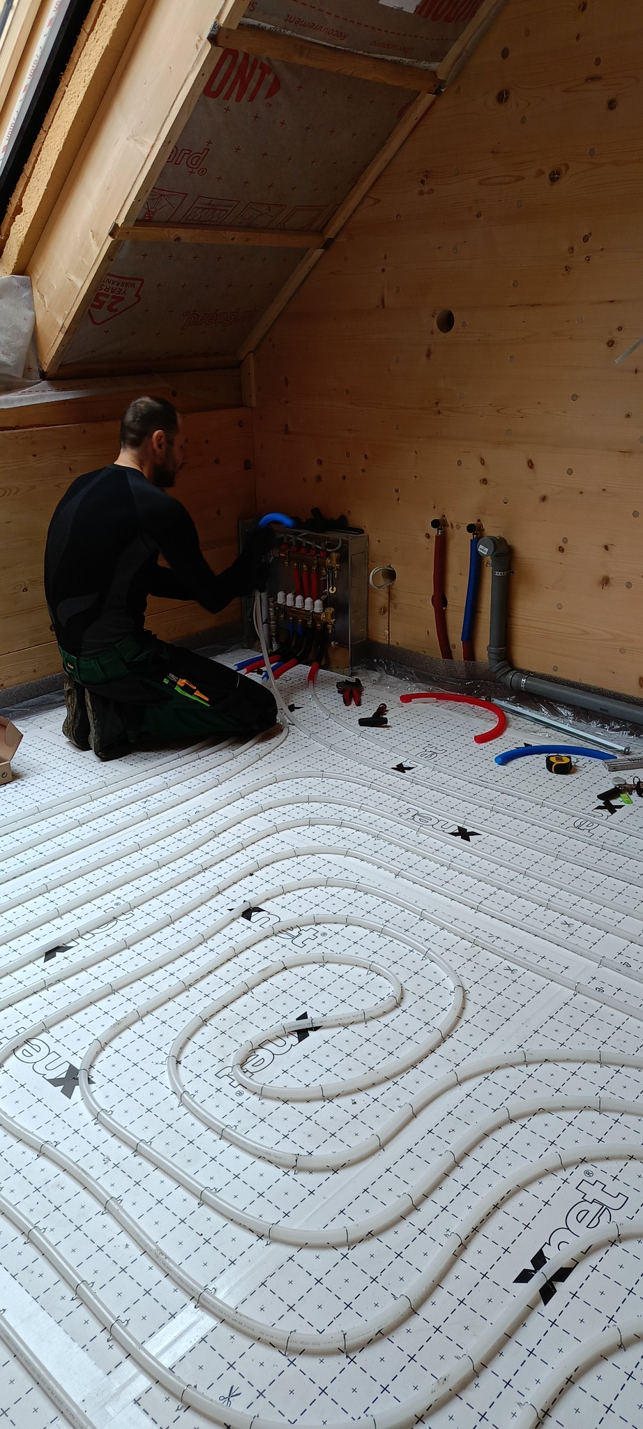 Instalacja ogrzewania podłogowego w drewnianym domu. Hydraulik łączy rury do rozdzielacza. Widoczne narzędzia i elementy instalacji. Ujęcie z góry, poddasze.