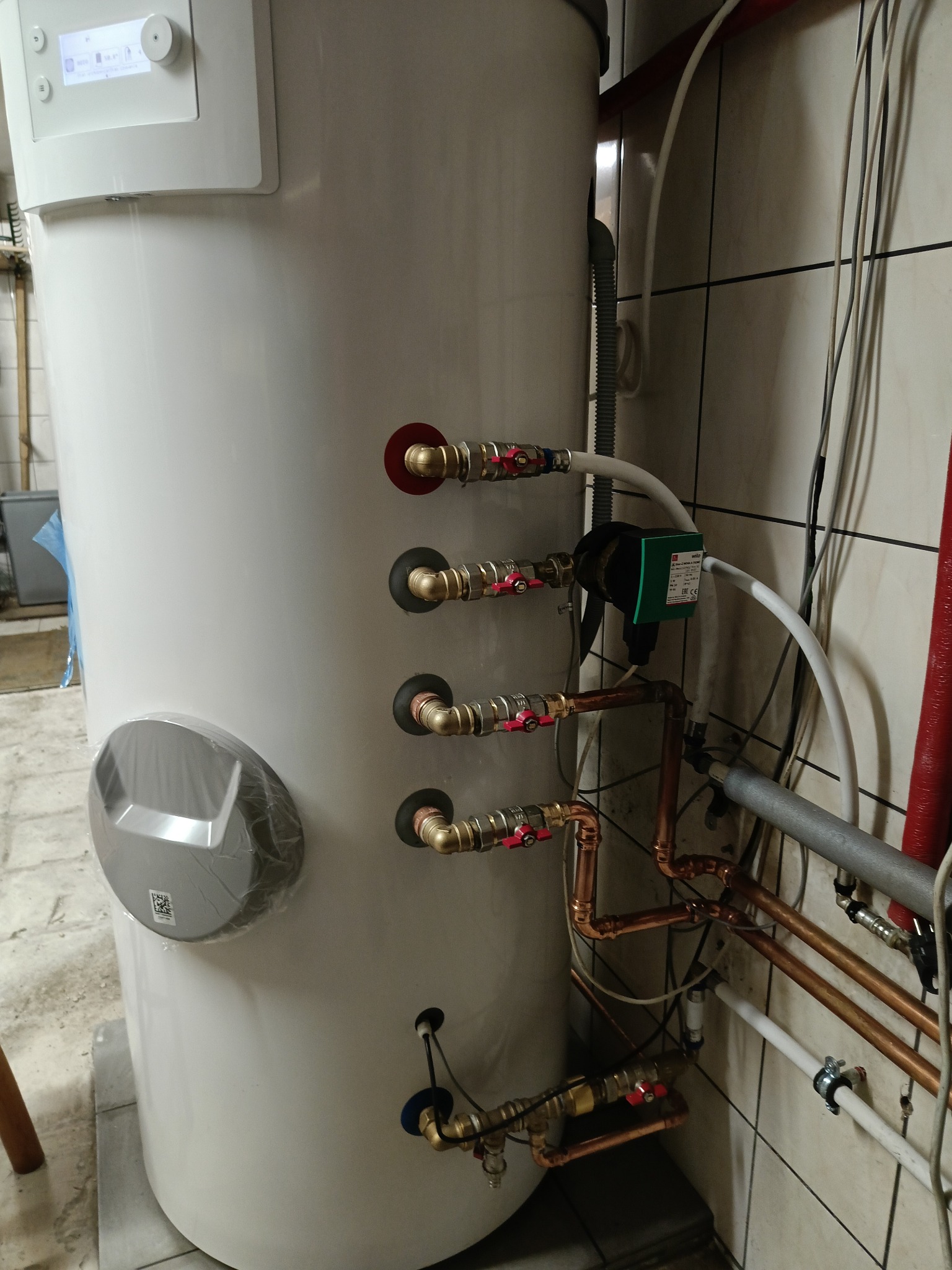 Biały zasobnik ciepłej wody użytkowej z licznymi przyłączami rur miedzianych i zaworów, zamontowany w pomieszczeniu z wykończeniem z płytek ceramicznych.
