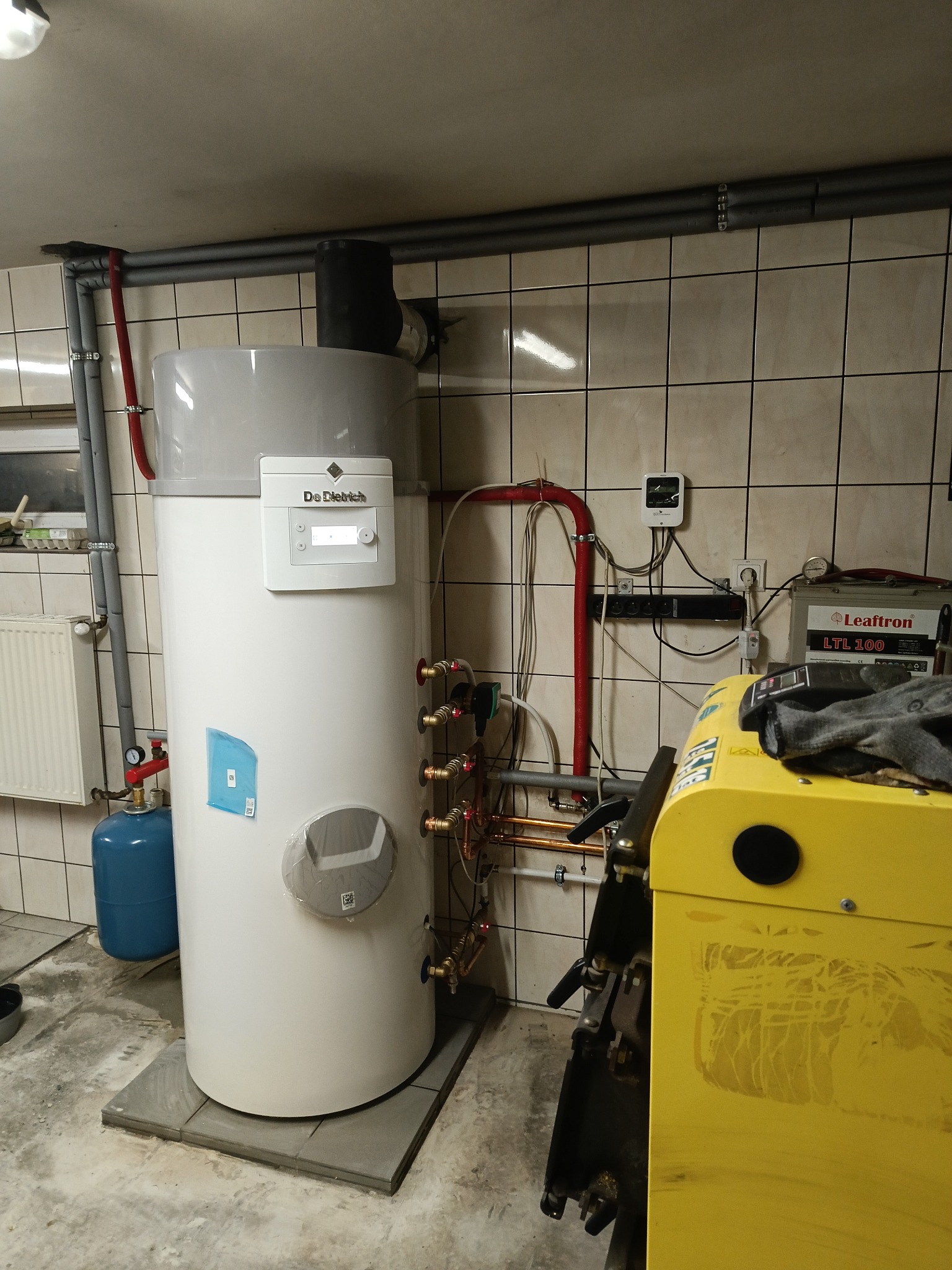 Biały piec gazowy De Dietrich z szarym zbiornikiem, zainstalowany w pomieszczeniu z kafelkami, widoczne rury i osprzęt, obok żółty sprzęt instalacyjny.