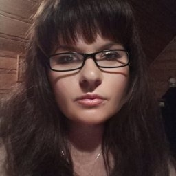 Beata Suchecka - Portret kobiety w okularach z ciemnymi włosami i grzywką, na tle drewnianej ściany. Widoczny delikatny łańcuszek na szyi. Zdjęcie z aplikacją FaceApp.