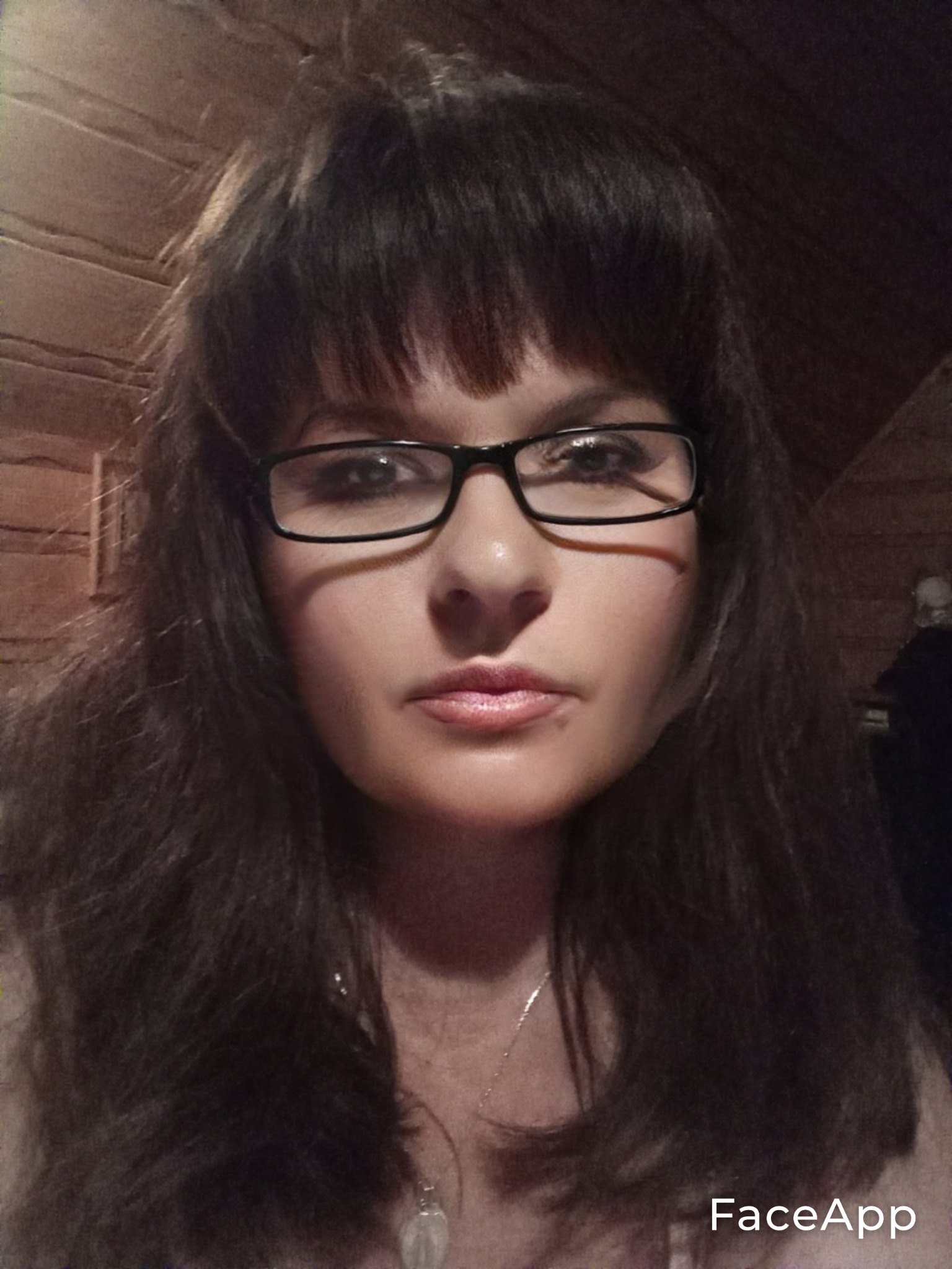 Portret kobiety w okularach z ciemnymi włosami i grzywką, na tle drewnianej ściany. Widoczny delikatny łańcuszek na szyi. Zdjęcie z aplikacją FaceApp.