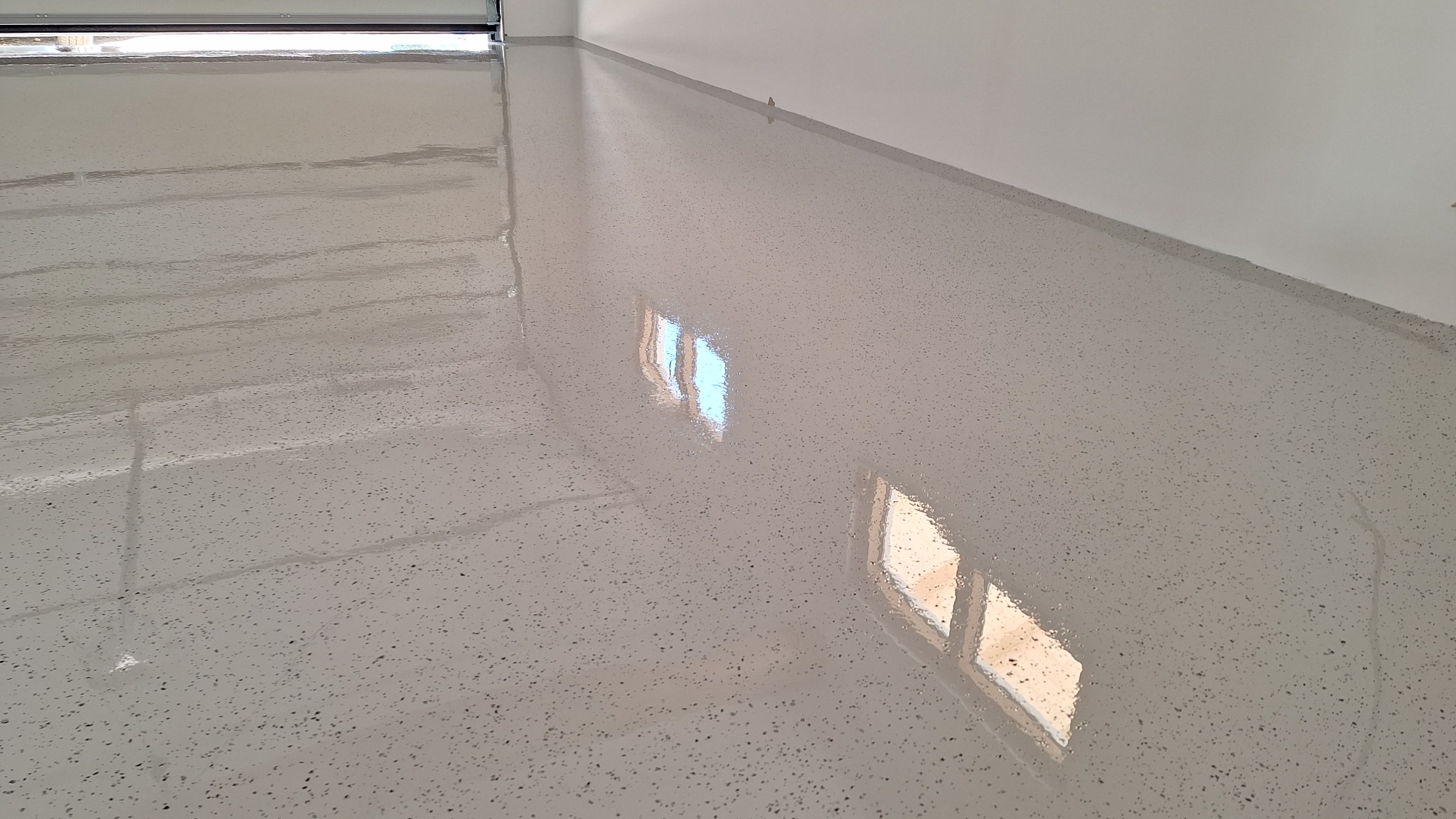 Jasnoszara, błyszcząca posadzka z żywicy z delikatnym wzorem drobnych, ciemnych kropek, odbijająca światło okna w garażu.