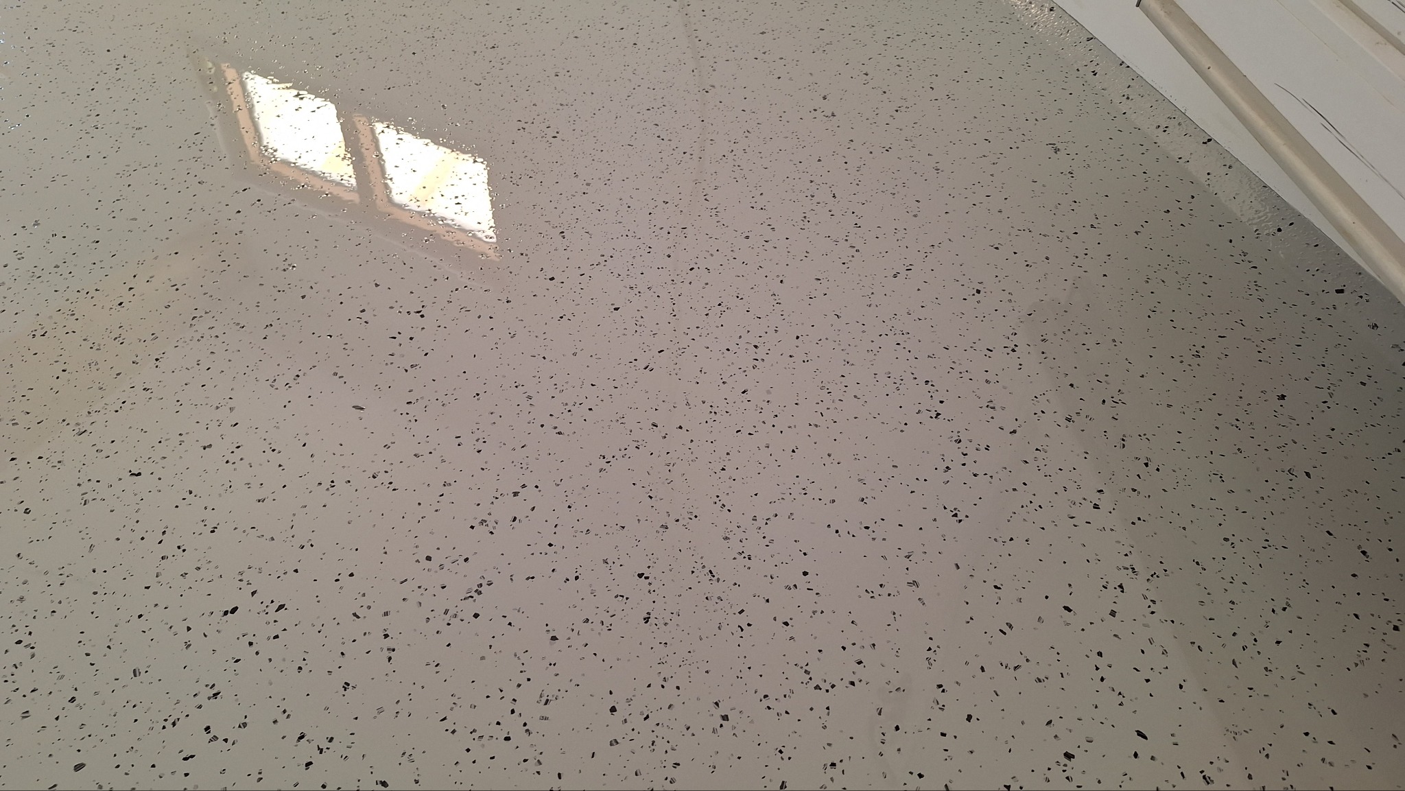 Jasnoszara posadzka żywiczna z czarnymi drobinami, odbijająca światło okna. Gładka powierzchnia z widoczną strukturą, fragment ściany w prawym górnym rogu.