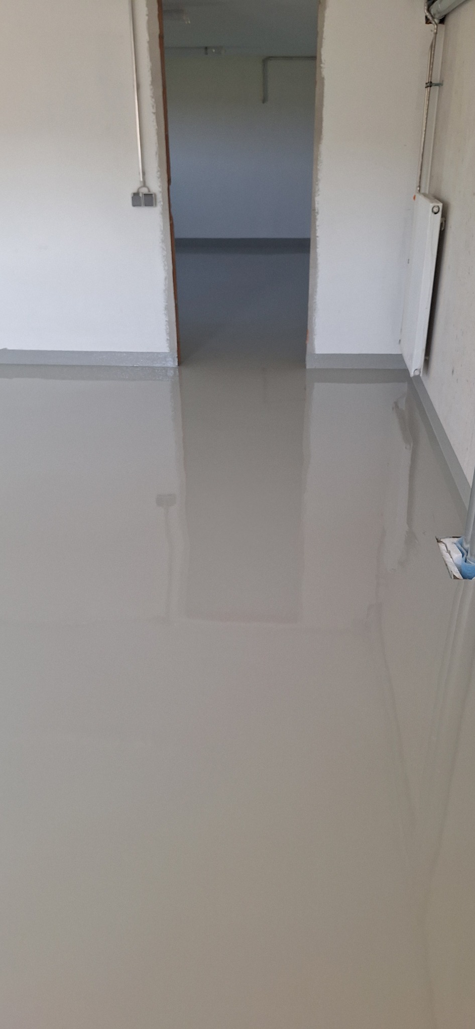 Jasnoszara, gładka posadzka w pomieszczeniu z białymi ścianami i grzejnikiem. Widoczne przejście do kolejnego pokoju z identyczną podłogą. Minimalistyczne wykończenie.