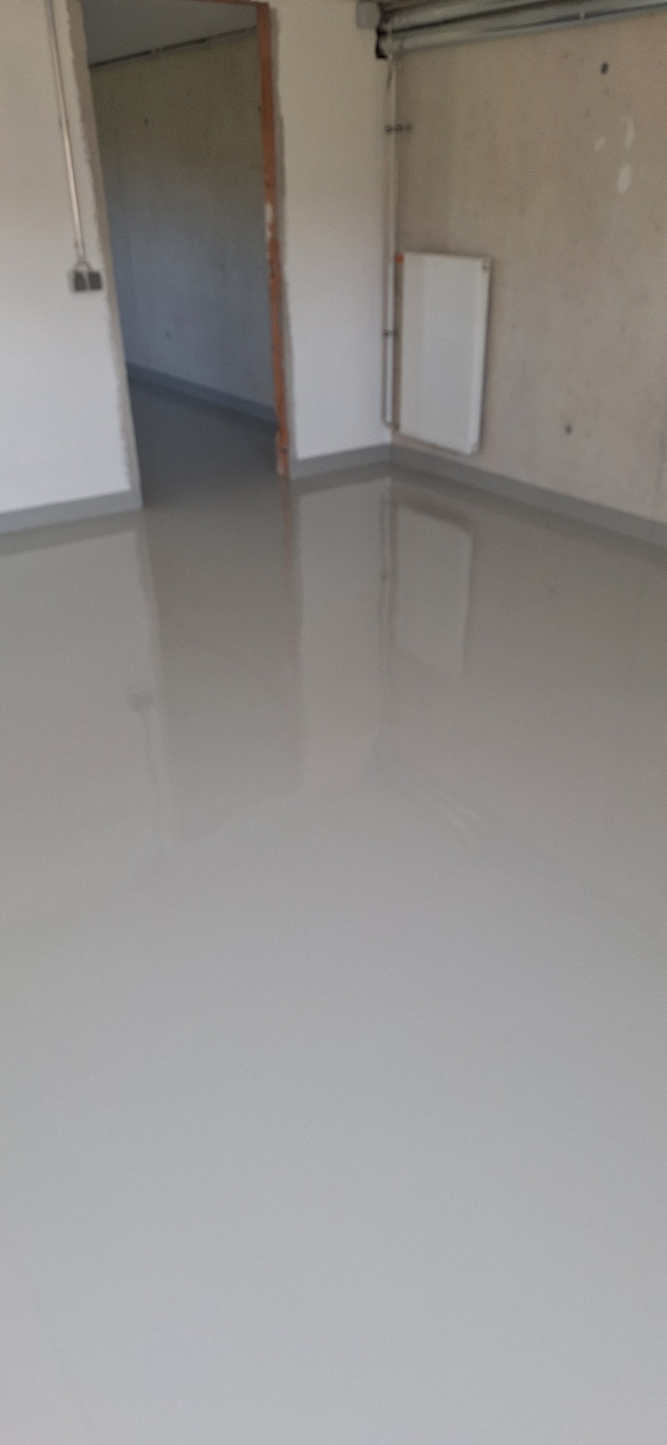 Jasnoszara, gładka posadzka w surowym wnętrzu z grzejnikiem na ścianie i otwartym przejściem do innego pomieszczenia. Widoczna refleksja światła na powierzchni.