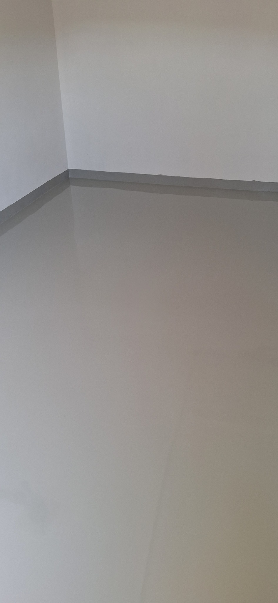 Gładka, szara posadzka w rogu pomieszczenia, z widoczną listwą przypodłogową. Minimalistyczne wnętrze z jednolitą powierzchnią podłogi.