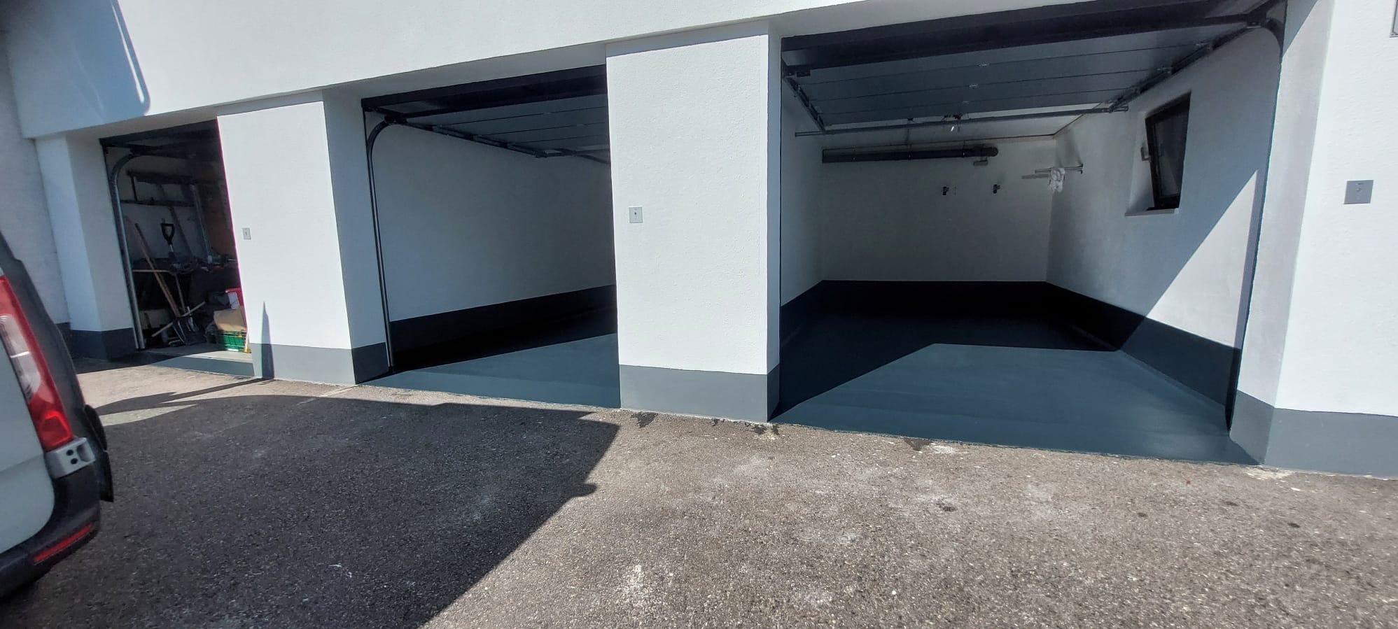 Gładka, szara posadzka w trzech garażach z otwartymi bramami, widok z zewnątrz. Jeden garaż z narzędziami w środku. Świeżo pomalowane ściany na biało.