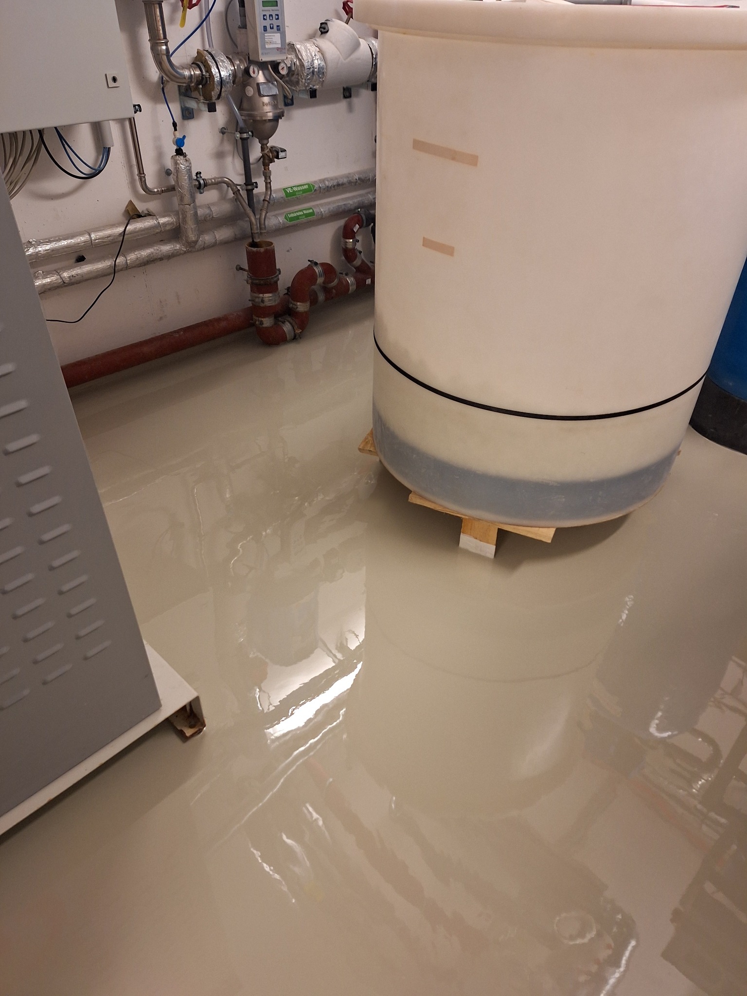 Błyszcząca, beżowa powierzchnia w pomieszczeniu technicznym z instalacjami i zbiornikiem. Widoczne odbicia w gładkiej powierzchni.