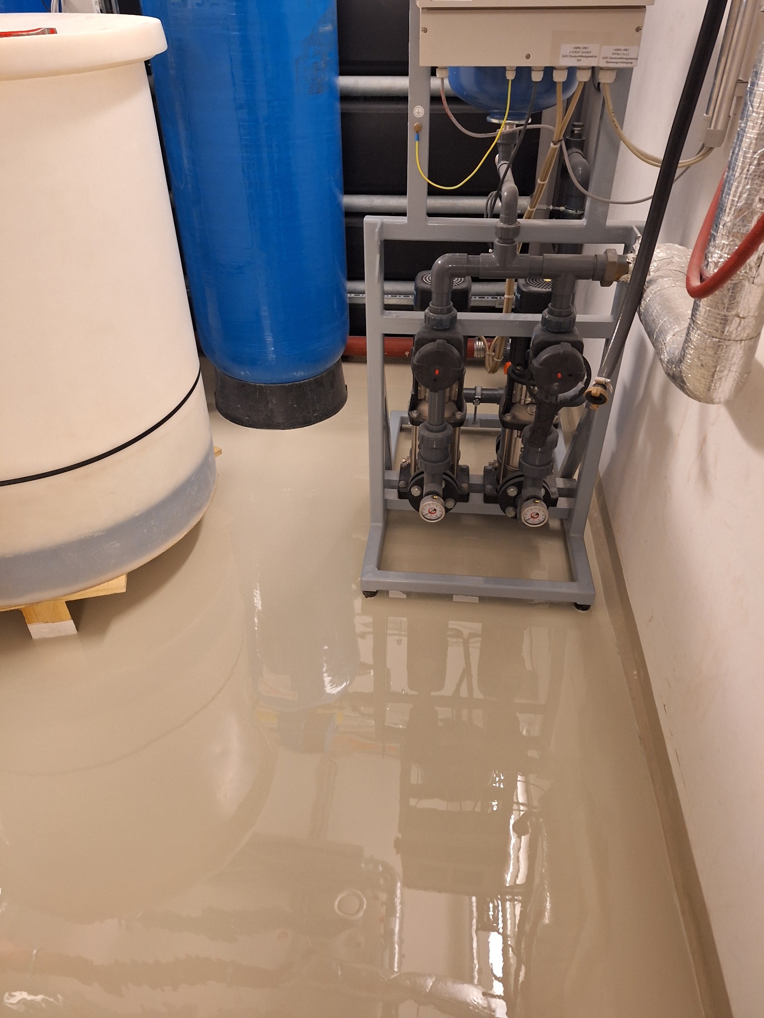 Jasna, błyszcząca posadzka w pomieszczeniu technicznym z instalacjami. Widoczne zbiorniki i system rur. Gładka powierzchnia podłogi odbija światło.