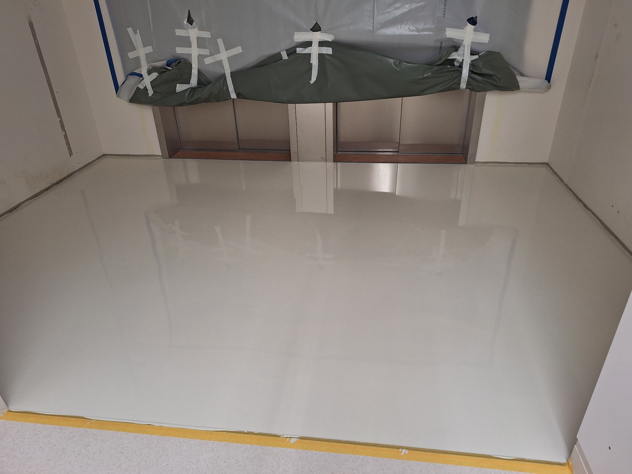 Jasna, gładka posadzka w pomieszczeniu z zabezpieczonym wejściem do windy. Żółta taśma maskująca przy ścianie. Widoczne odbicie światła na powierzchni.