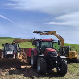 Sławomir Dziubdziela - Praca ziemne w toku: koparka CAT i ciągnik CASE przy wykopie na tle zielonego wzgórza i błękitnego nieba z smugami kondensacyjnymi.