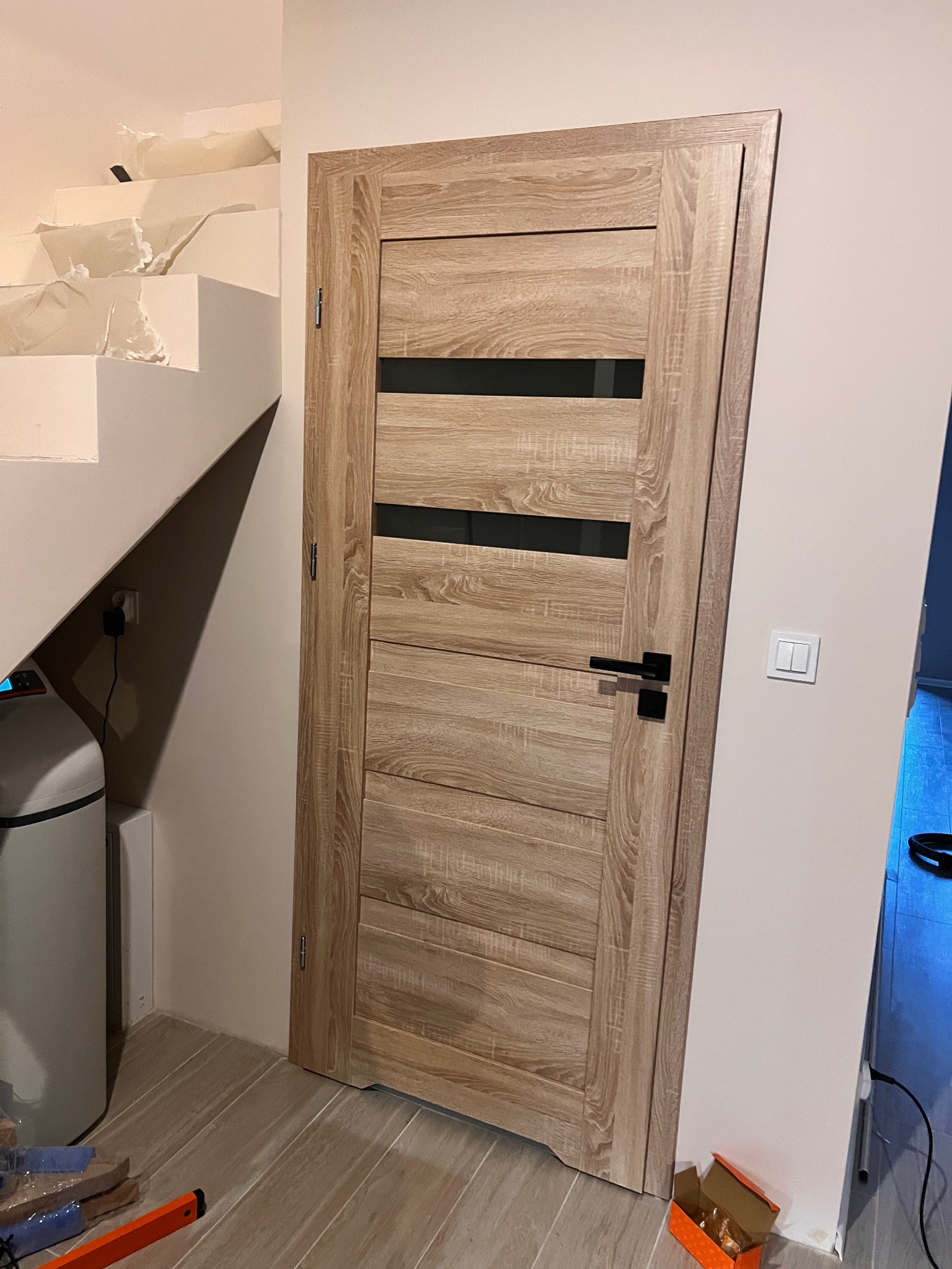 Zamknięte, drewniane drzwi z czarnymi, poziomymi przeszkleniami, zamontowane w jasnym wnętrzu. Widoczne schody w trakcie wykończenia i akcesoria montażowe.