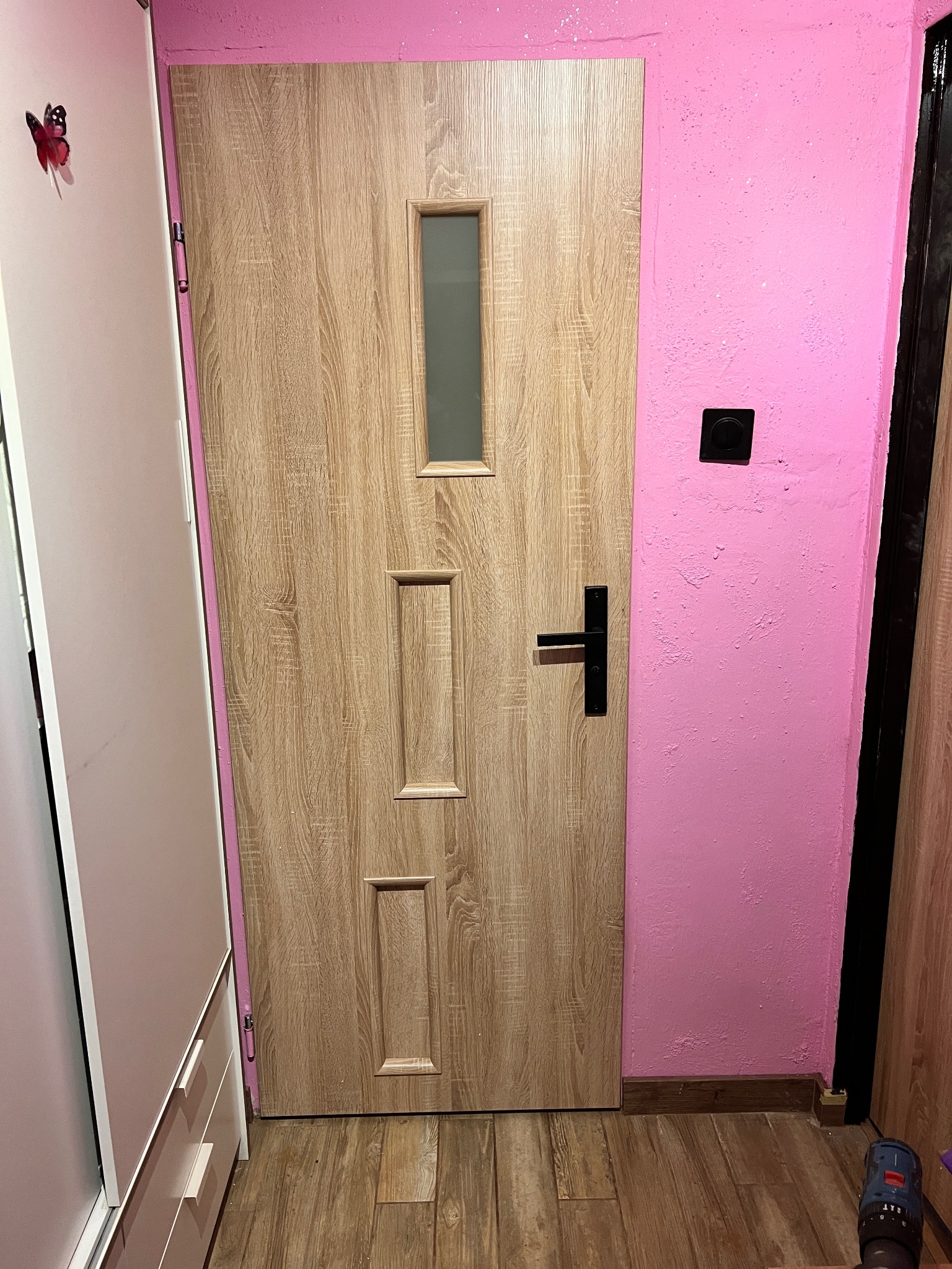 Zamontowane drzwi wewnętrzne z szybką i czarną klamką, widoczne na tle różowej ściany i drewnianej podłogi. Obok szafa z motylem na drzwiach.