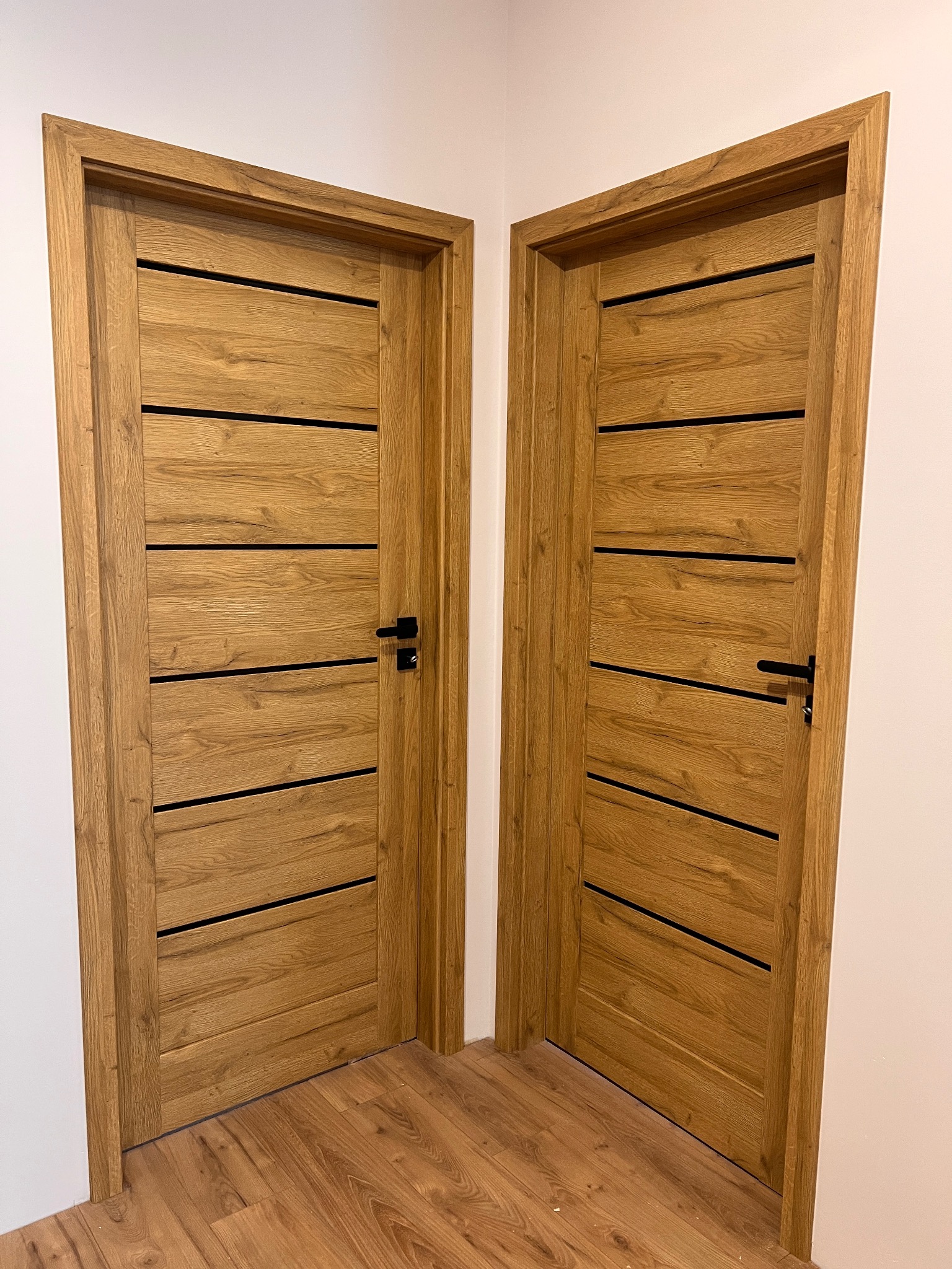 Dwa drewniane, nowoczesne drzwi wewnętrzne z czarnymi, poziomymi wstawkami, zamontowane w rogu pomieszczenia z panelami podłogowymi o podobnym odcieniu drewna.