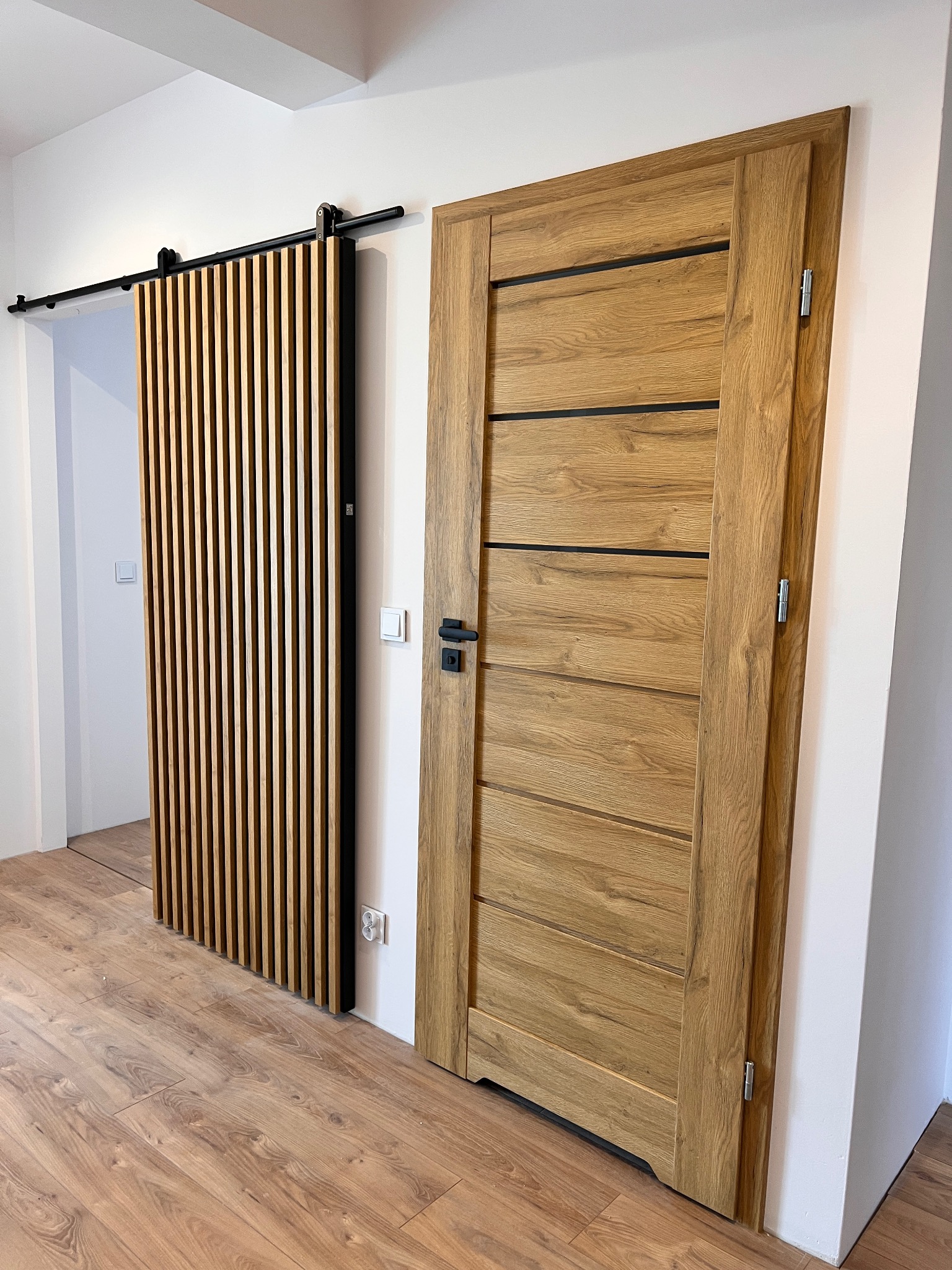 Dwie drewniane drzwi: klasyczne z czarnymi akcentami i przesuwne lamelowe na czarnej szynie. Jasne wnętrze, drewniana podłoga, minimalistyczny design.