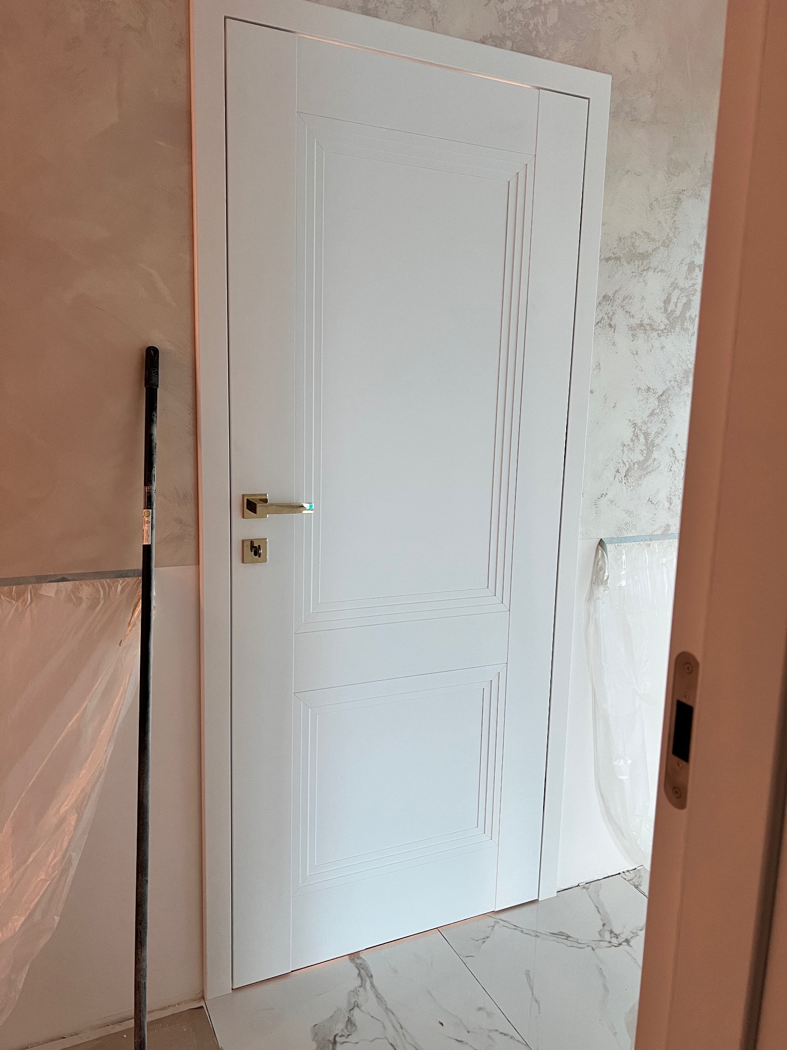 Białe, panelowe drzwi z złotą klamką, zamontowane w jasnym wnętrzu z marmurową podłogą. Widoczny fragment ściany z dekoracyjnym tynkiem i zabezpieczenia folią malarską.