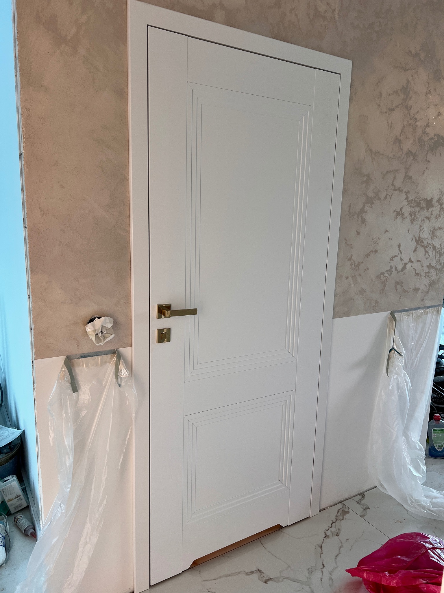 Białe, panelowe drzwi z złotą klamką, zamontowane w odnowionym wnętrzu. Widoczne zabezpieczenia folią malarską i fragment ściany z dekoracyjnym tynkiem.