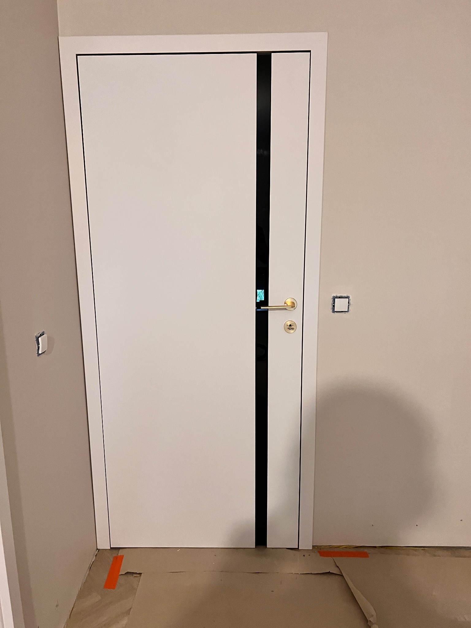 Białe drzwi z czarnym pionowym pasem i złotą klamką, zamontowane w jasnym wnętrzu. Widoczne ślady po remoncie na podłodze i puszka elektryczna na ścianie.