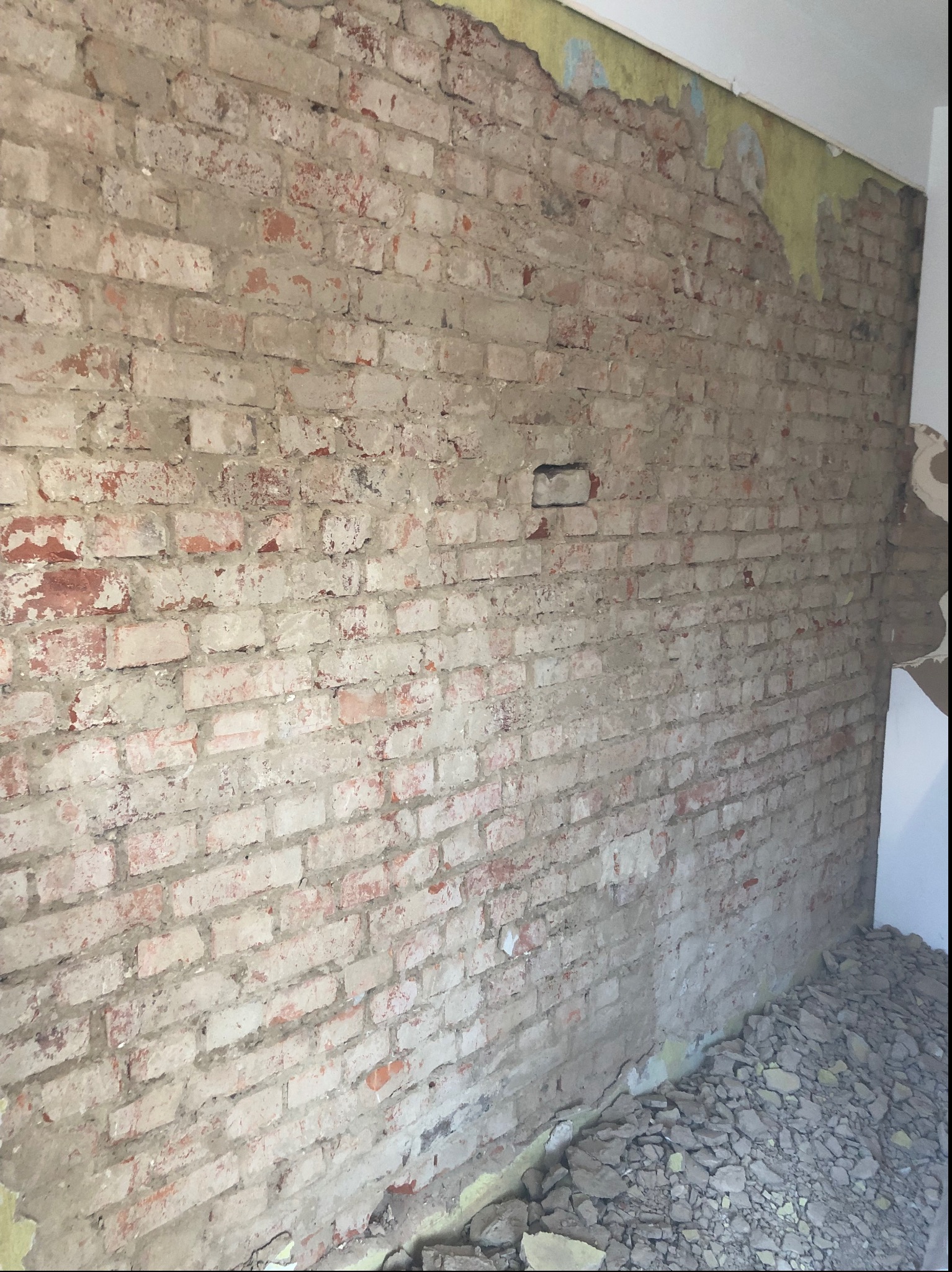 Odsłonięta ceglana ściana w trakcie prac rozbiórkowych, z widocznymi resztkami tynku i gruzem u podstawy. Fragment ściany z żółtą farbą.