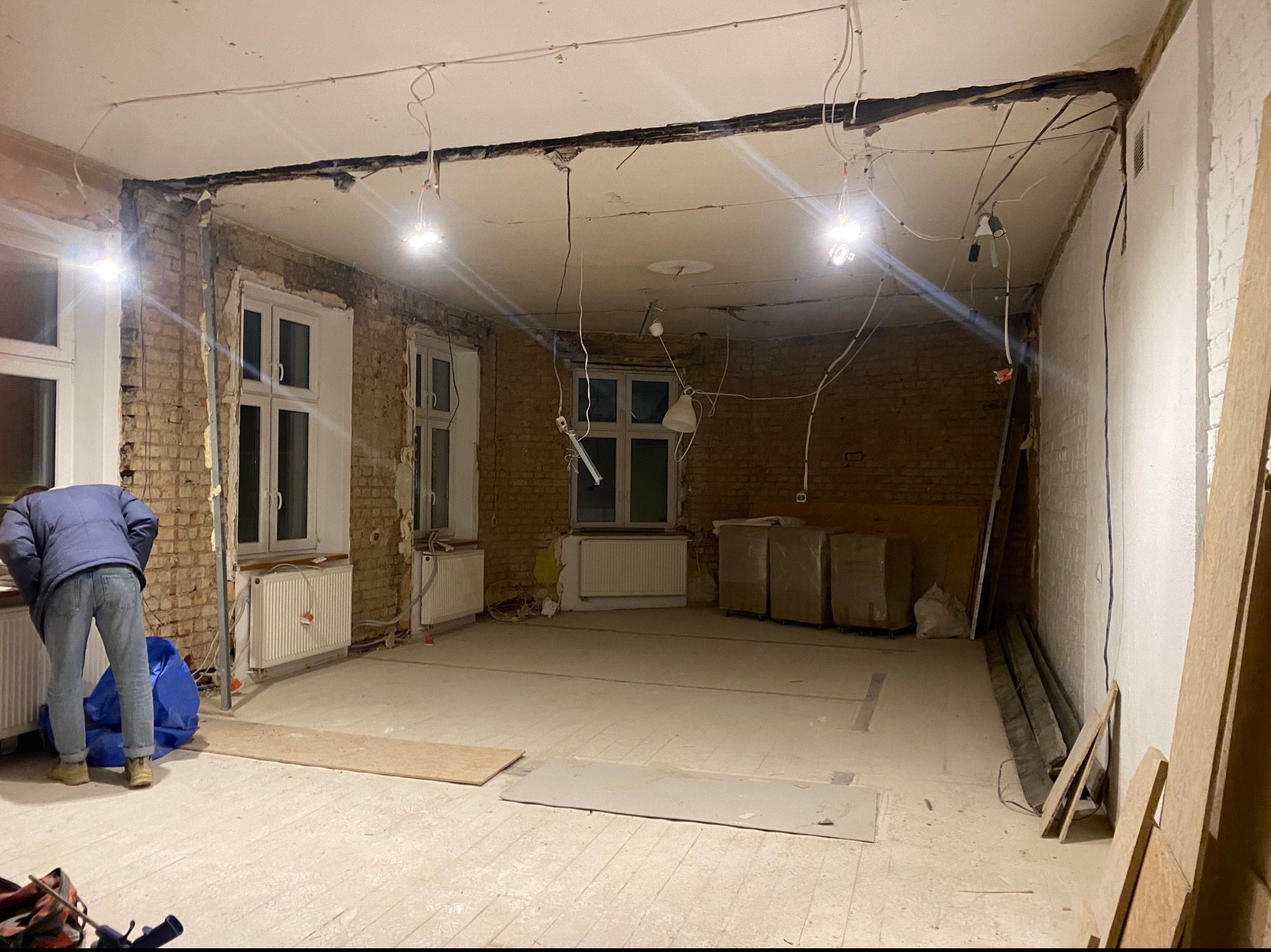 Wnętrze po demontażu ścian i sufitu podwieszanego. Widoczne odsłonięte cegły, instalacje i fragment osoby w trakcie prac porządkowych. Oświetlenie sufitowe.