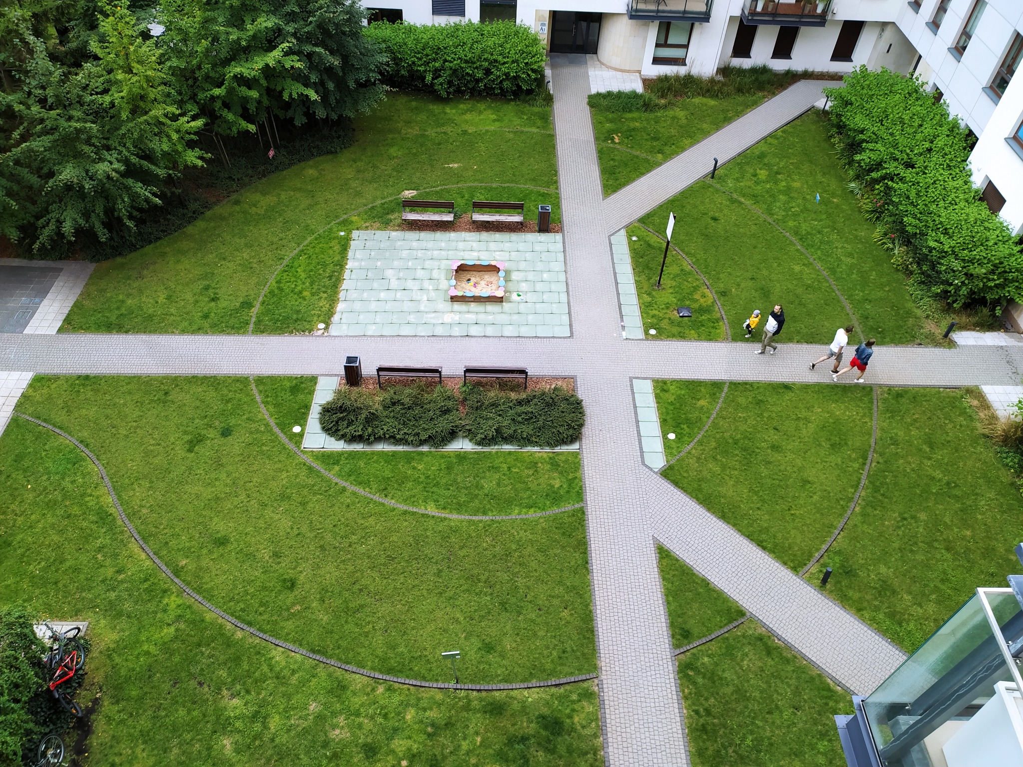 Widok z góry na zadbany dziedziniec osiedla z zielonym trawnikiem, alejkami z kostki, placem zabaw z piaskownicą i ławeczkami. W tle budynek mieszkalny.