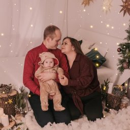 Fotografia Rodzinna Irina Kazarinova - Rodzinne zdjęcie w świątecznej scenerii: rodzice patrzą na siebie, trzymając ubrane w renifera dziecko na tle choinki i dekoracji.