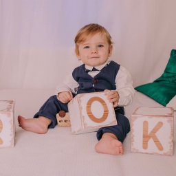 Fotografia Rodzinna Irina Kazarinova - Uroczy chłopiec w eleganckim stroju siedzi na łóżku, otoczony drewnianymi klockami z literami układającymi się w słowo 'ROK'. Jasne tło, miękkie światło.