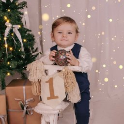 Fotografia Rodzinna Irina Kazarinova - Roczne dziecko w stroju galowym trzyma błyszczącą bombkę na tle choinki, prezentów i dekoracji urodzinowych. Urocza sesja w ciepłych barwach z efektem bokeh.